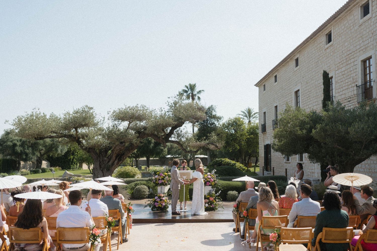 Outdoor wedding ceremony at a rustic venue with guests seated on wooden chairs surrounded by olive trees and a stone building.