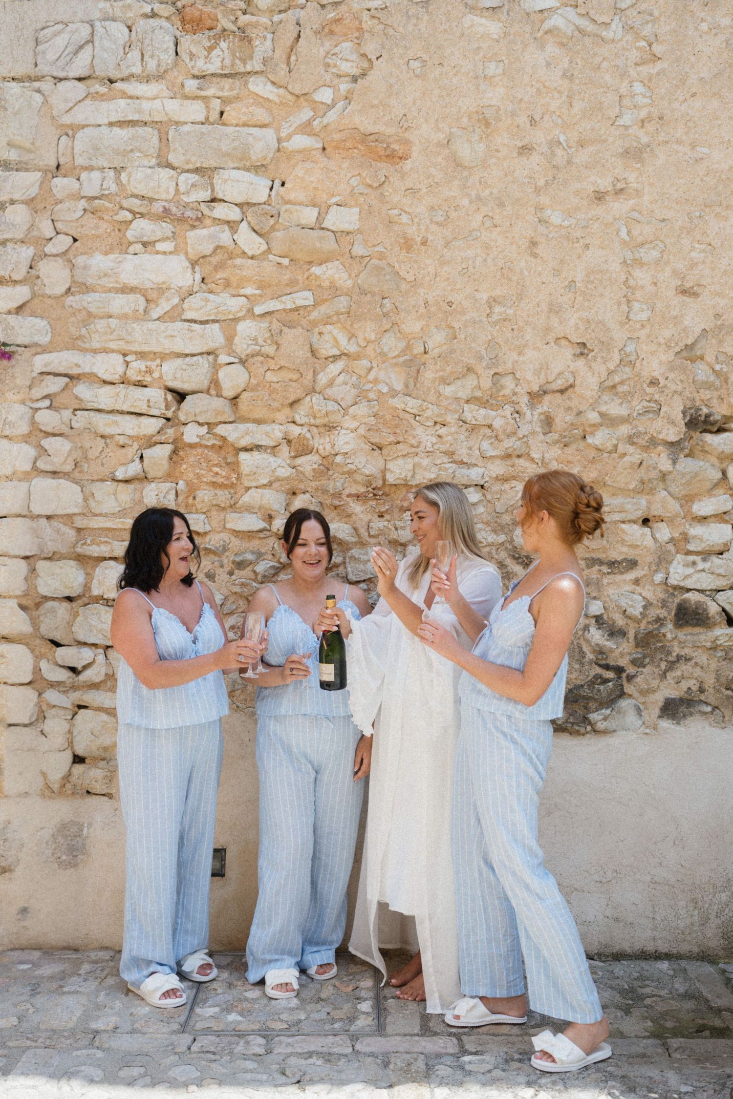 Four bridesmaids in blue striped pajamas celebrate with champagne against a rustic stone wall.