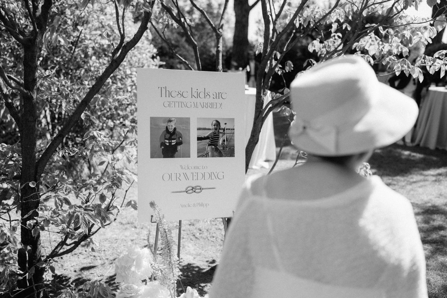 Black and white photo of a person in a white hat viewing an outdoor wedding sign display among trees.
