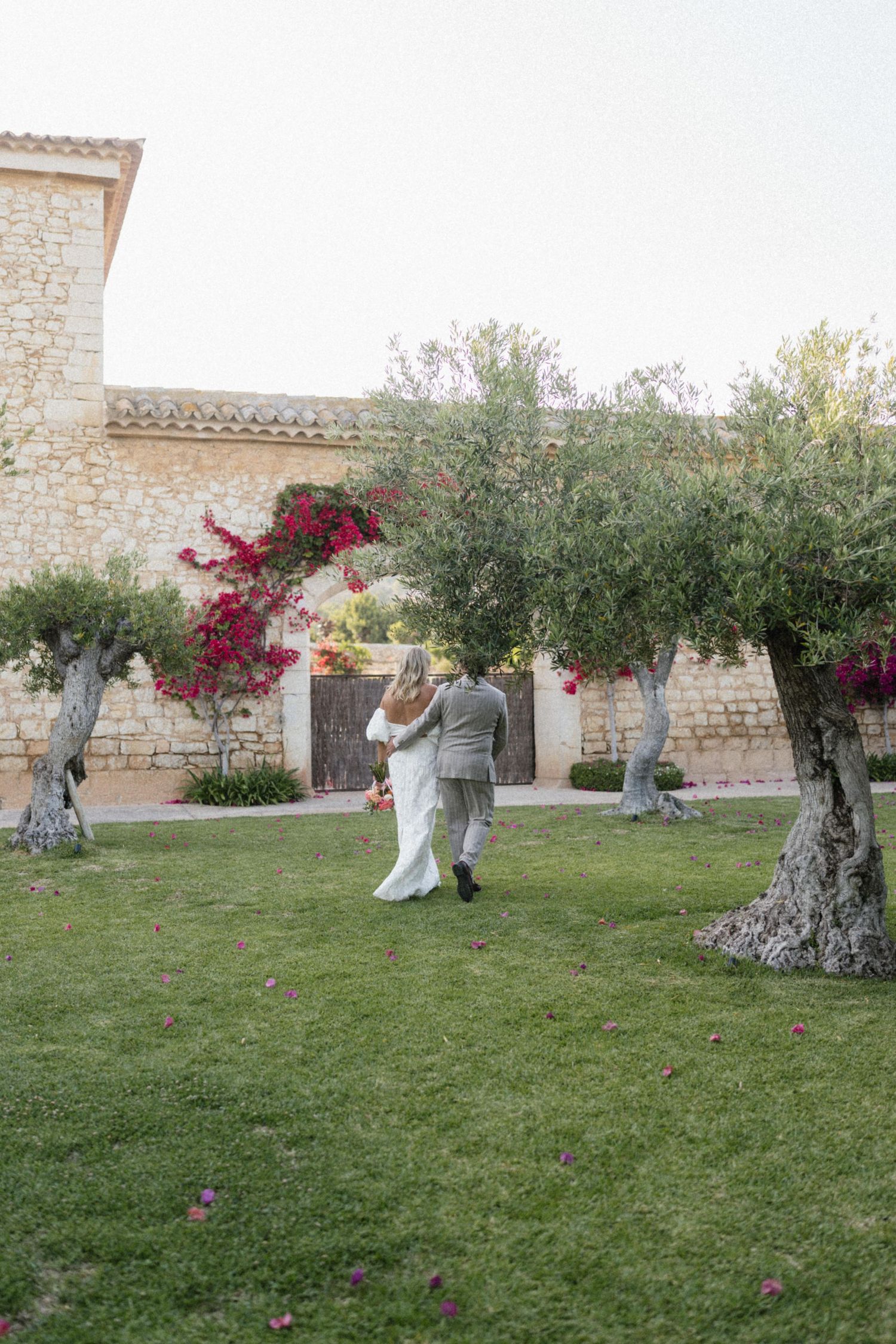 Bride and groom dancing on lush green lawn beside olive trees with bougainvillea at a Mediterranean stone villa wedding.