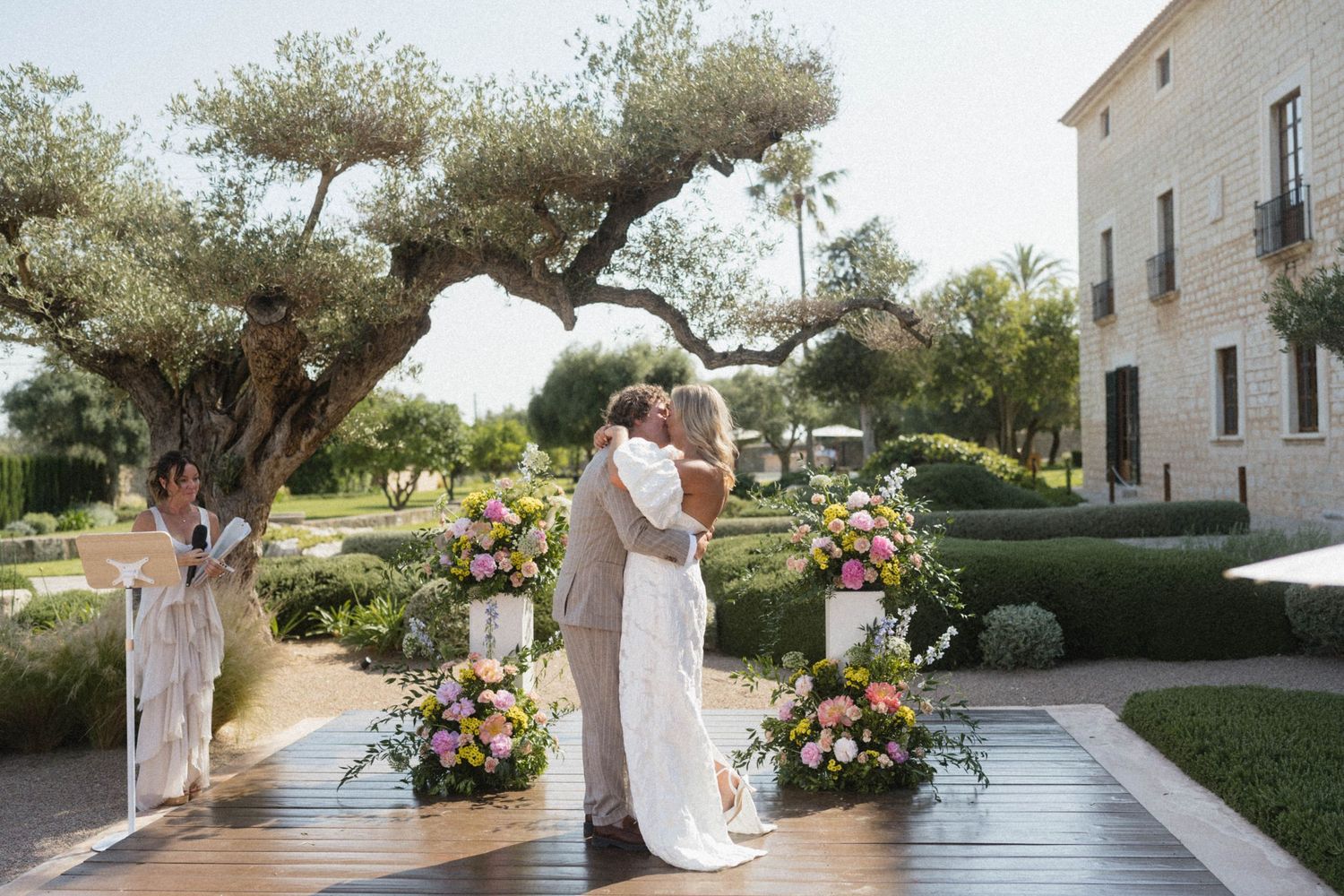 Bride and groom share first kiss at elegant outdoor wedding ceremony with colorful floral arrangements.