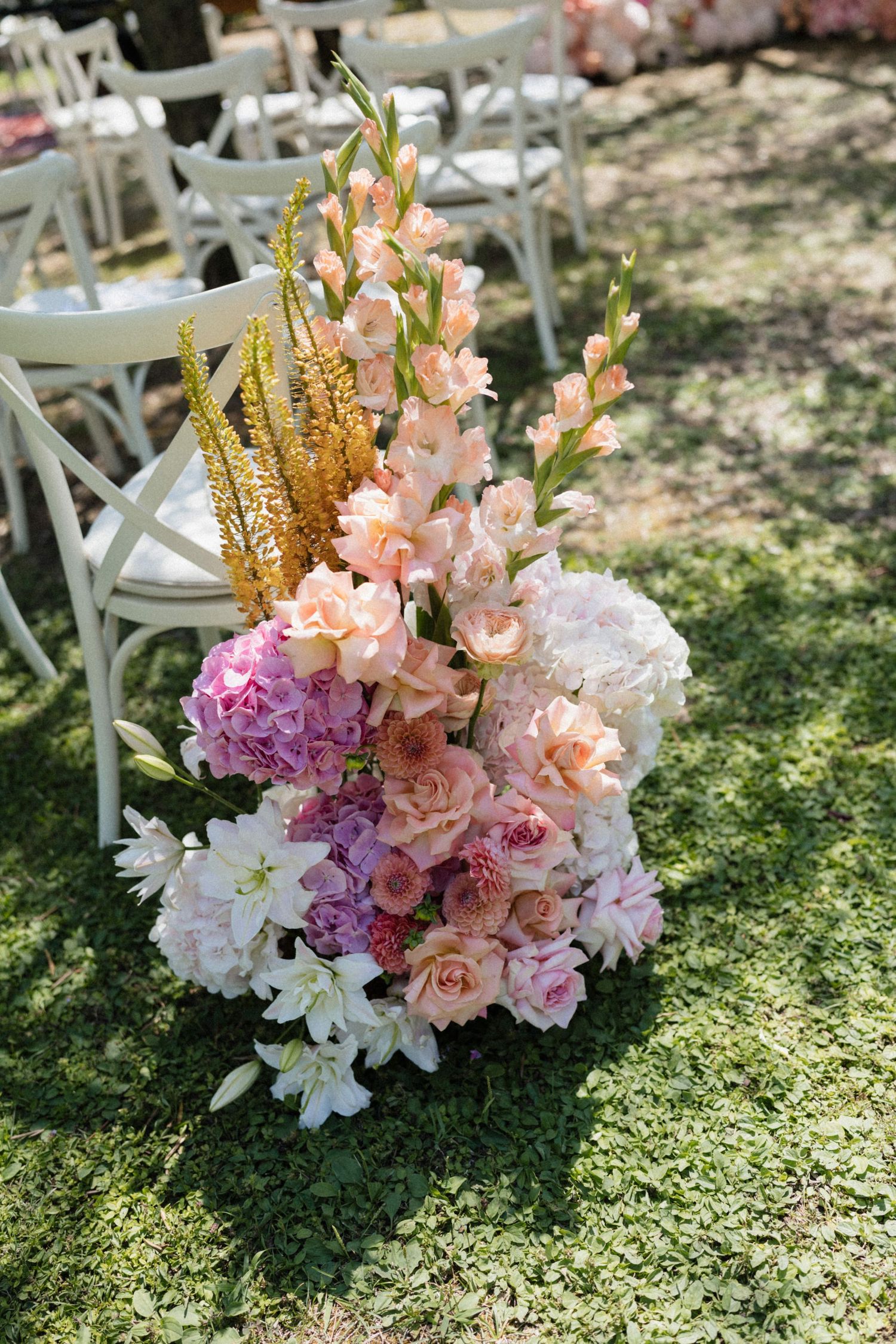 Lush outdoor wedding ceremony aisle floral arrangement with pink hydrangeas, roses, gladiolus, and gold accents on green grass.
