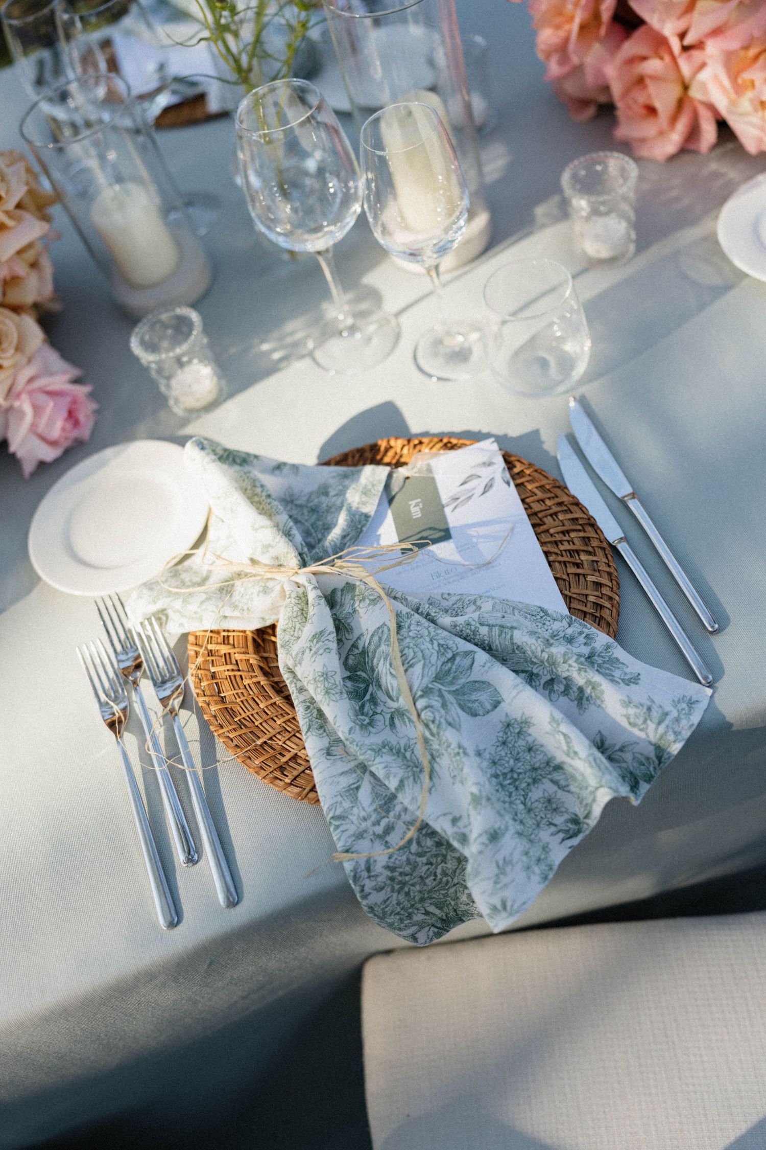 Elegant wedding table setting with wicker charger, floral napkin, silverware, crystal glasses, and pink roses on a light blue tablecloth.