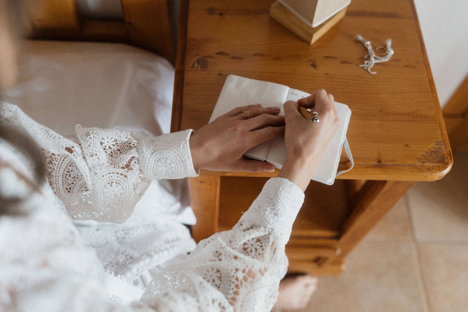 Bride in lace dress writing in journal at wooden nightstand on wedding day.