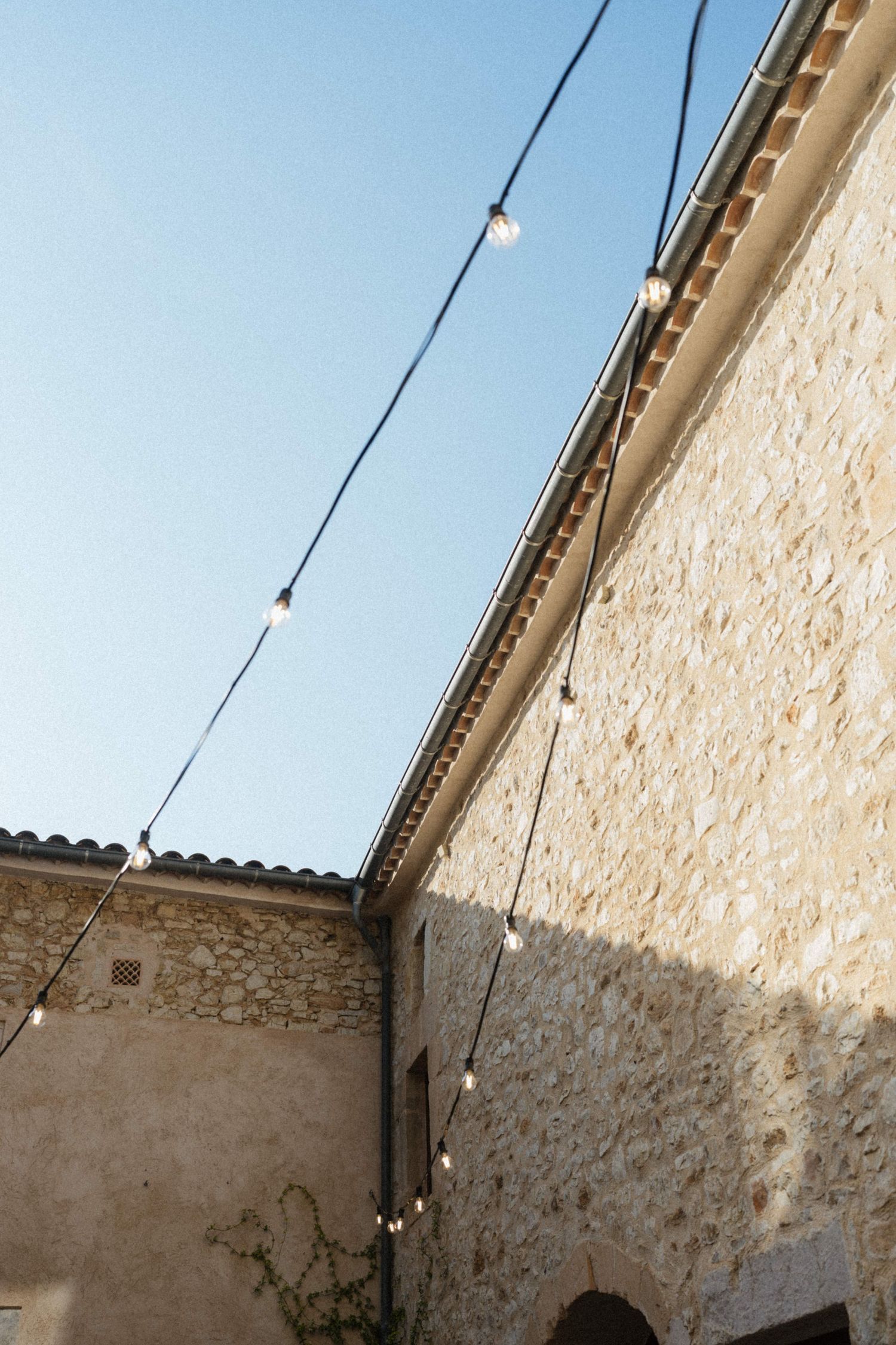 String lights hang between rustic stone buildings against a clear blue sky at an outdoor wedding venue.
