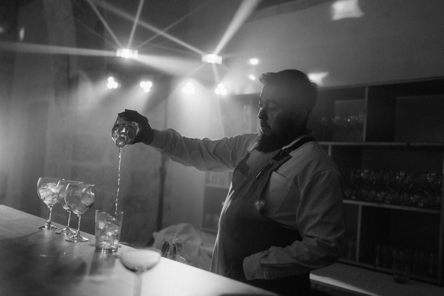 Bartender pouring drinks at a bar with dramatic stage lighting in black and white photo.