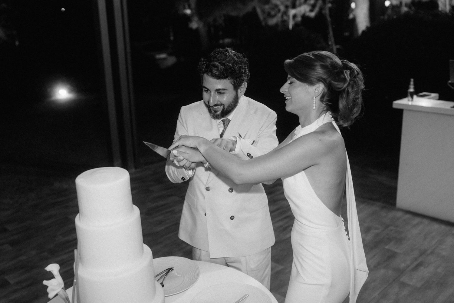 Black and white photo of bride and groom cutting wedding cake together at evening reception.