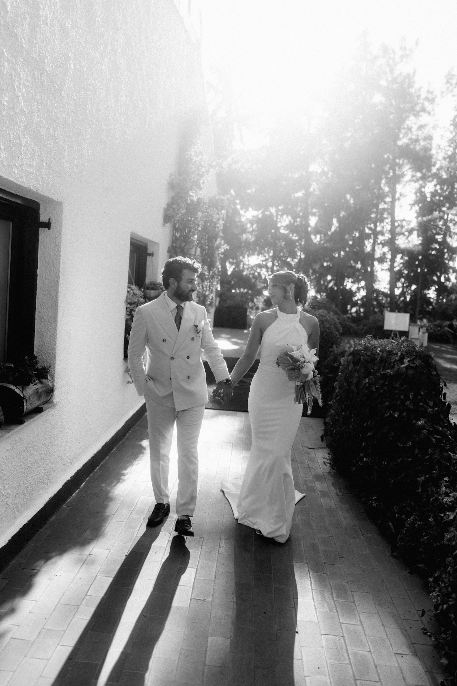 Black and white photo of a bride and groom holding hands, walking together in sunlight outside a white building.