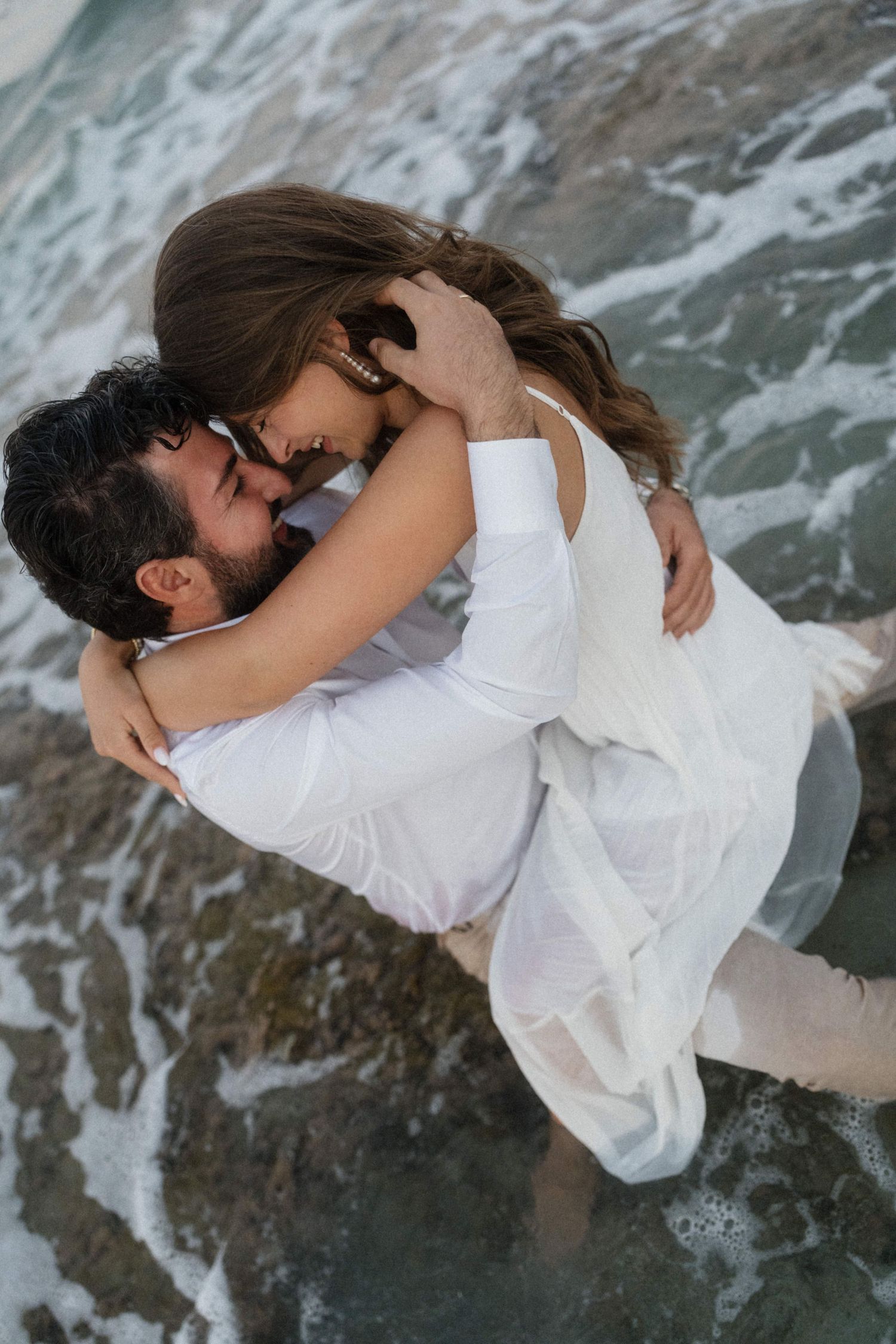 A couple in white clothing shares a romantic kiss while standing in ocean waves on a beach.