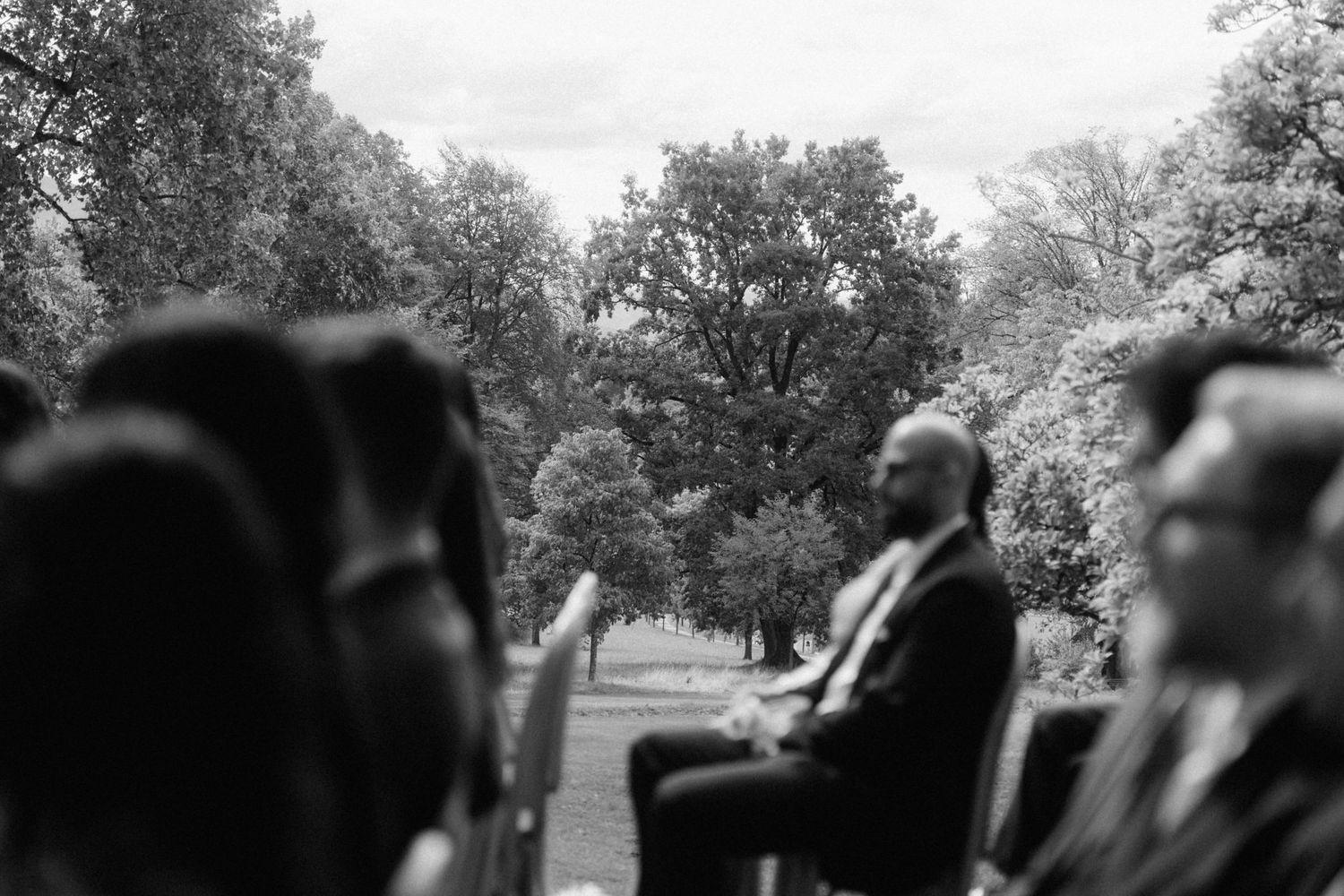 Black and white candid photo of wedding guests seated outdoors during garden ceremony among trees.