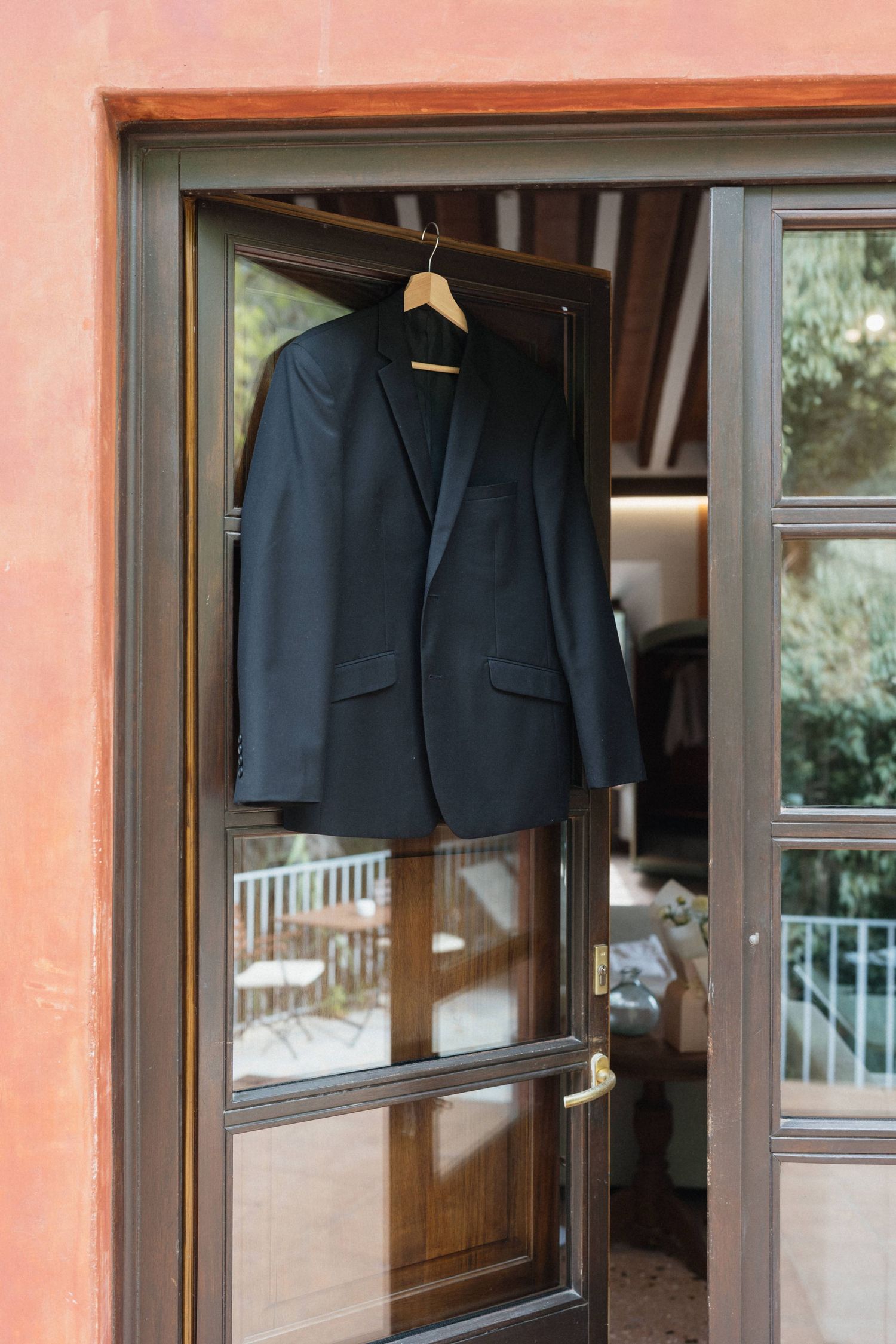 Dark suit jacket on a wooden hanger hanging from a brown-framed glass door of a terracotta building, with a sunlit patio and trees beyond.