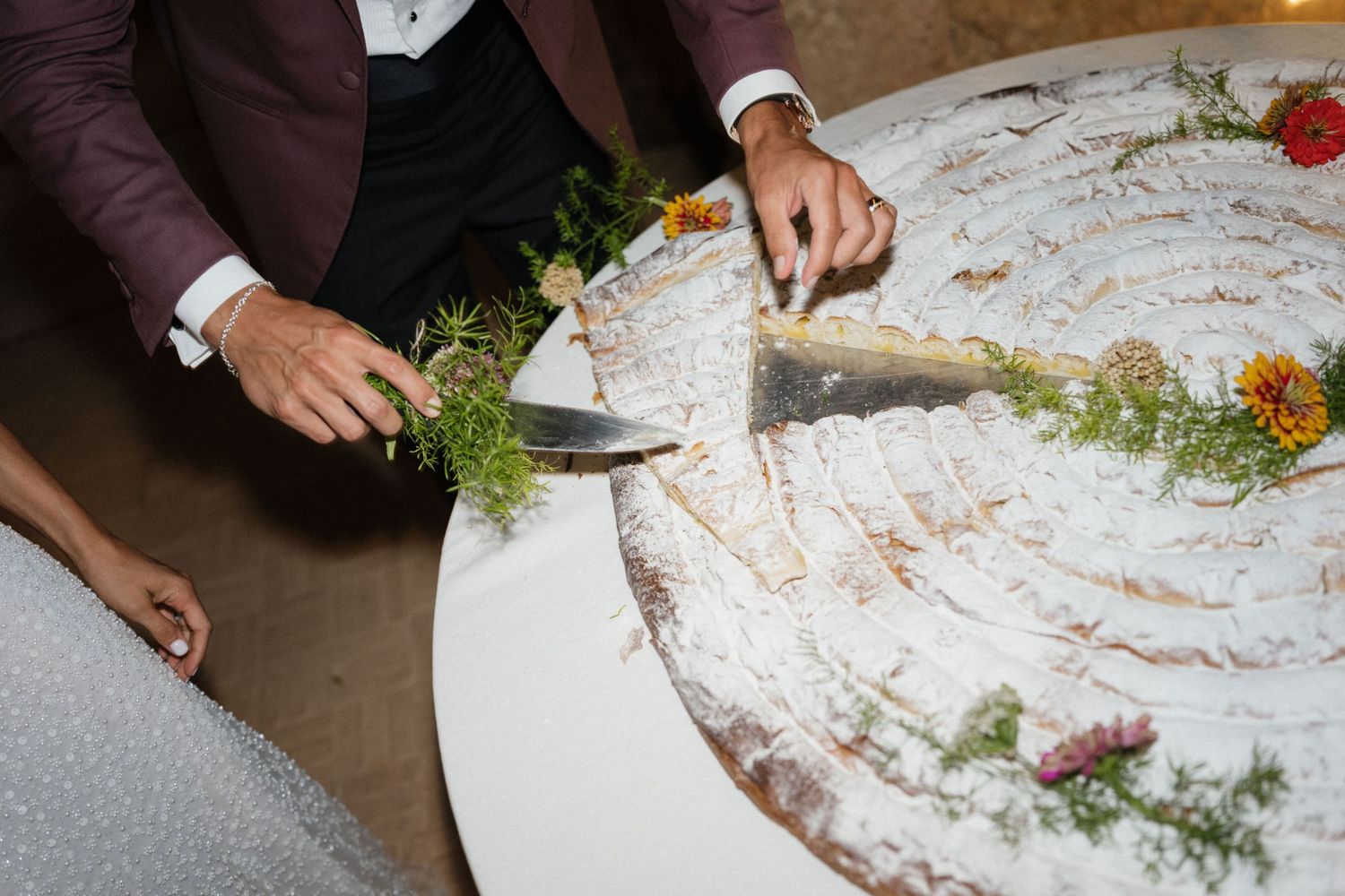 A groom in a burgundy jacket slices a large decorated wedding cake garnished with herbs and edible flowers.