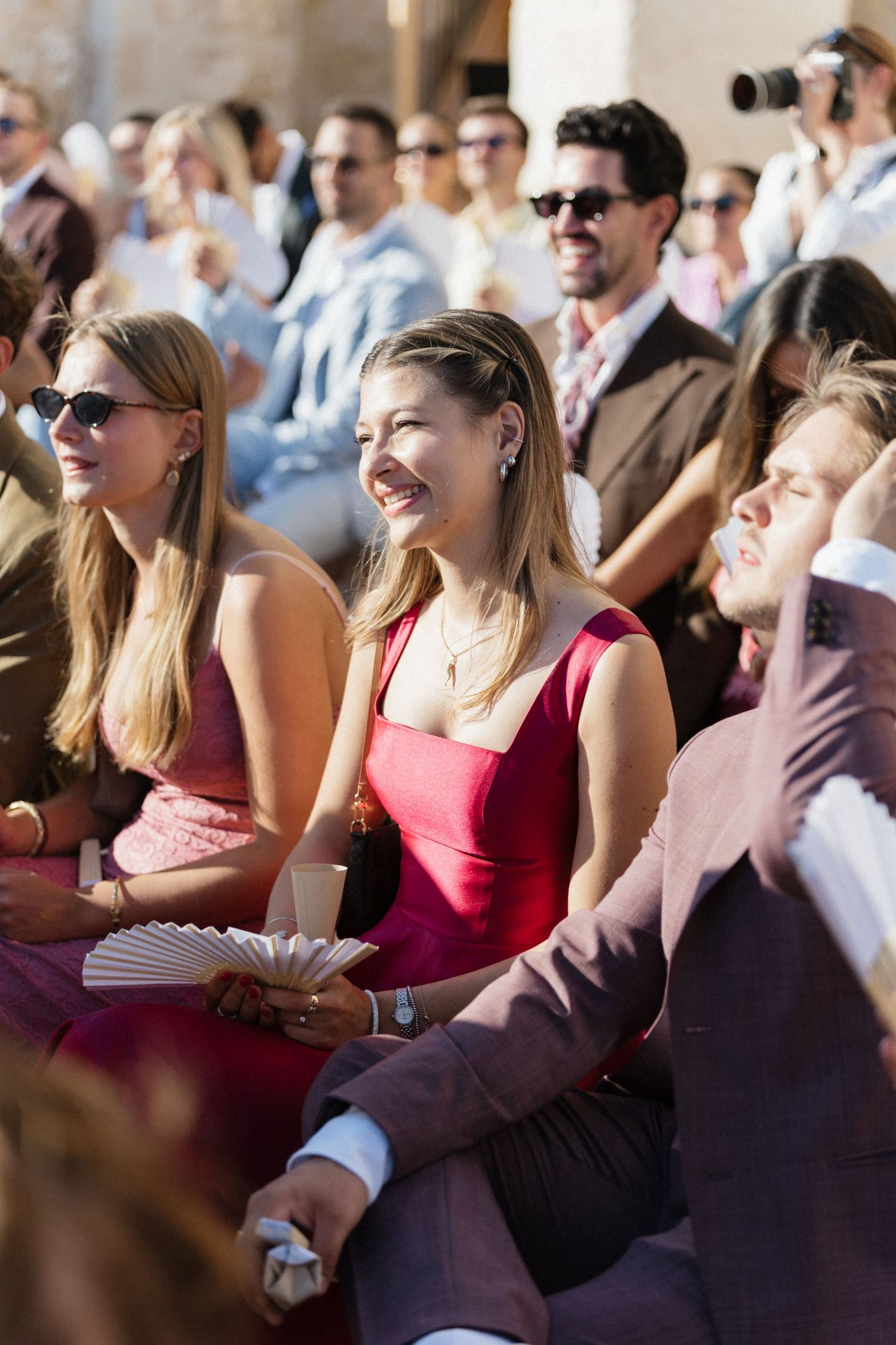 Smiling wedding guests in summer outfits sit close together at an outdoor ceremony, holding folding hand fans.