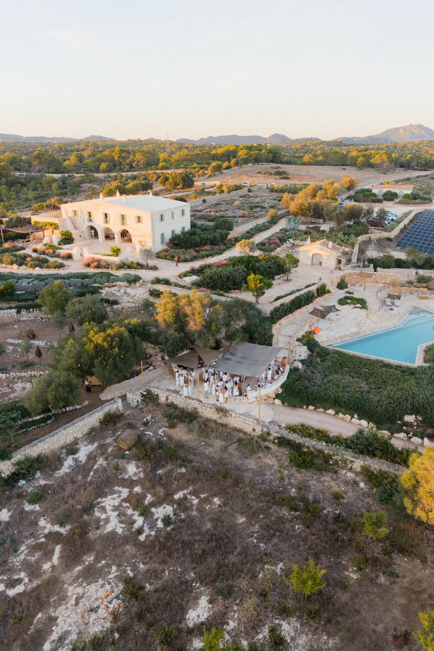 Aerial view of a whitewashed Mediterranean villa with a pool, olive groves and a wedding gathering on a stone terrace at sunset.