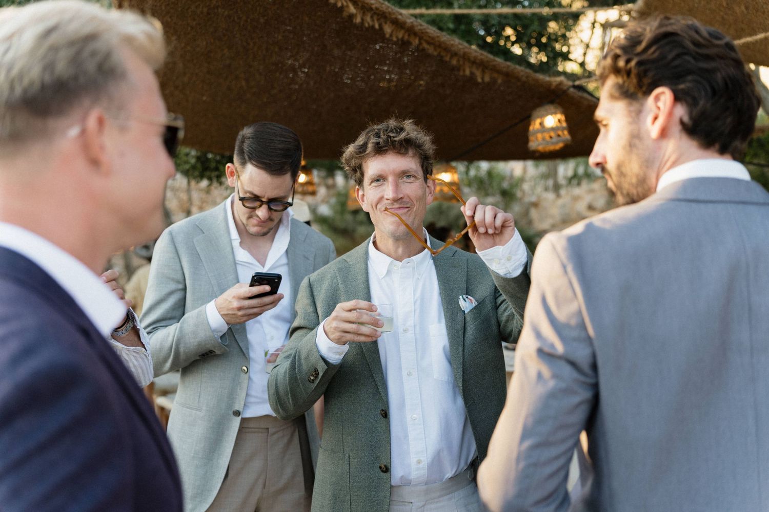 Three groomsmen in suits chat and laugh at an outdoor wedding reception while another checks his phone.