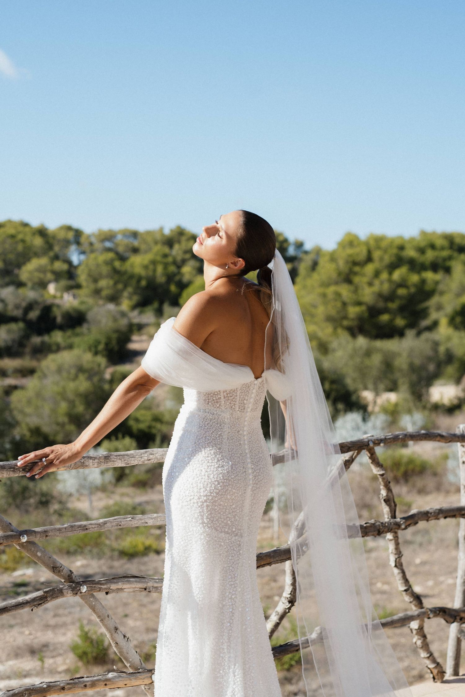 Bride in a sparkling off-shoulder wedding gown and long veil gazes skyward beside a rustic wooden railing.