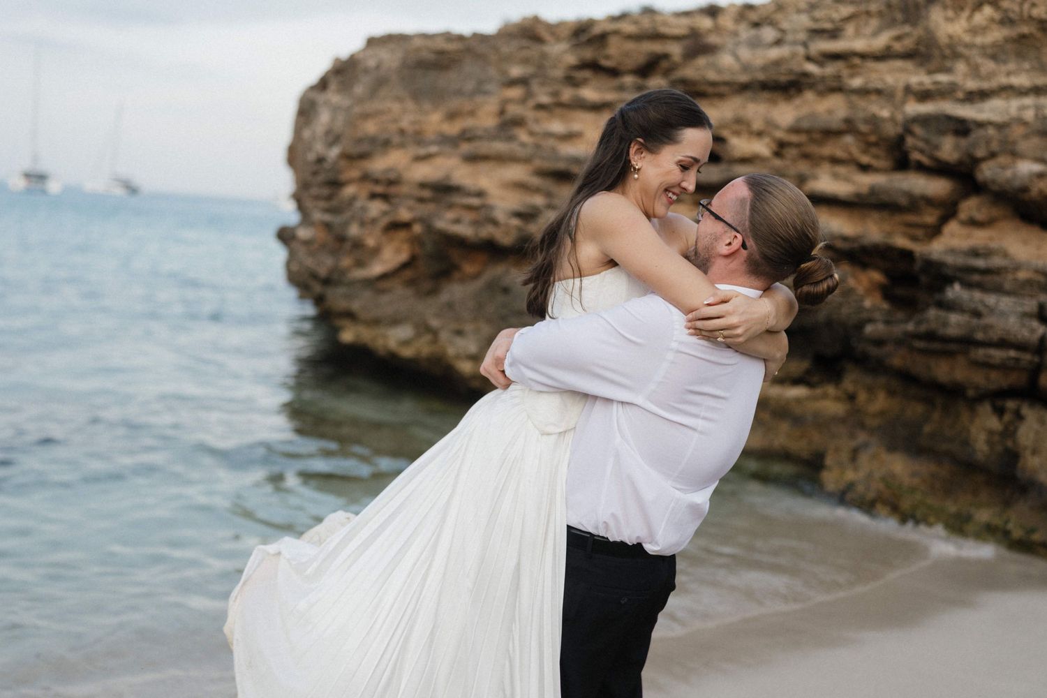 Groom in a white shirt lifts his smiling bride in a flowing white gown on a beach beside rocky cliffs and calm sea.