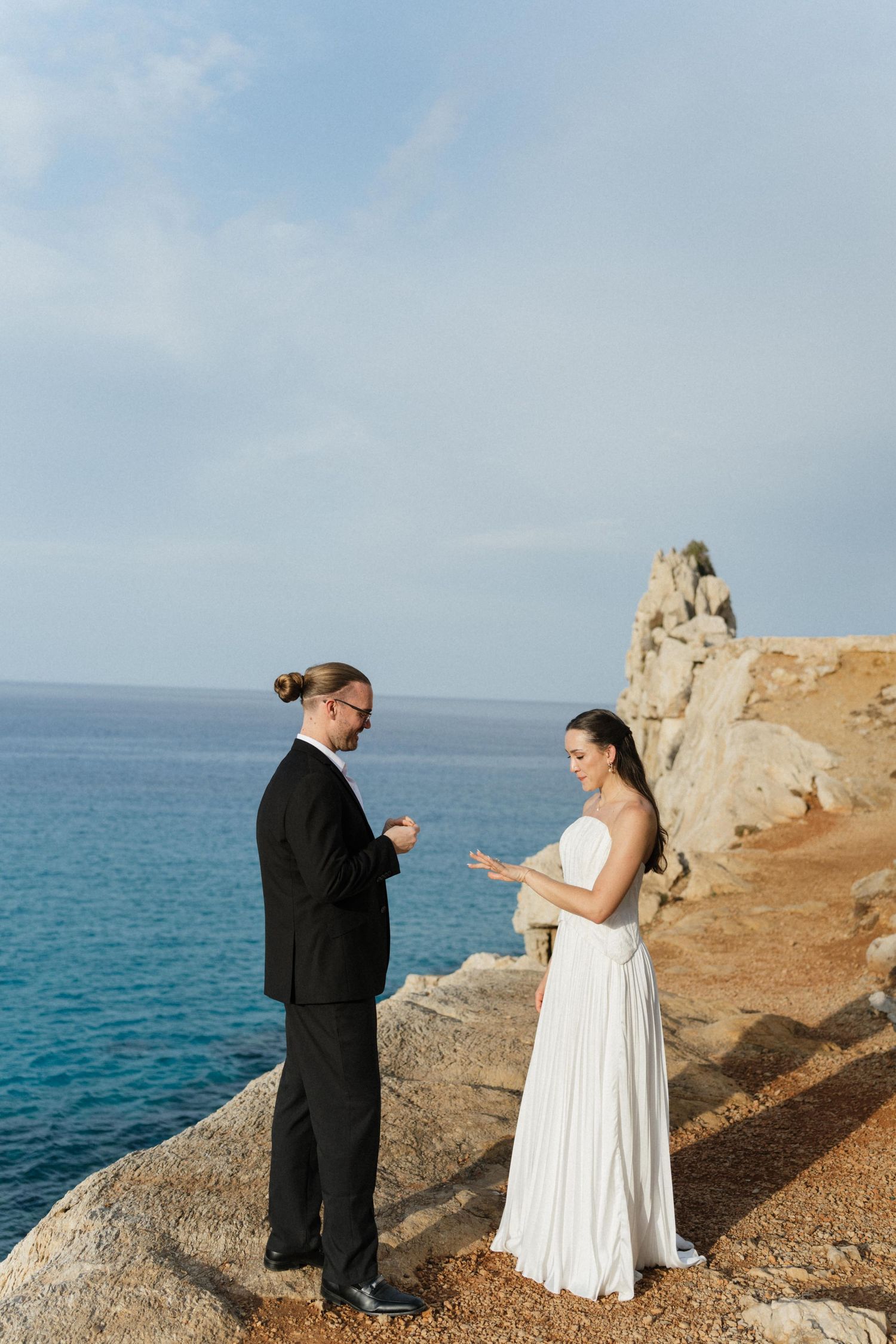 A bride and groom exchange rings on a rocky coastal cliff overlooking the blue sea during an intimate elopement ceremony.