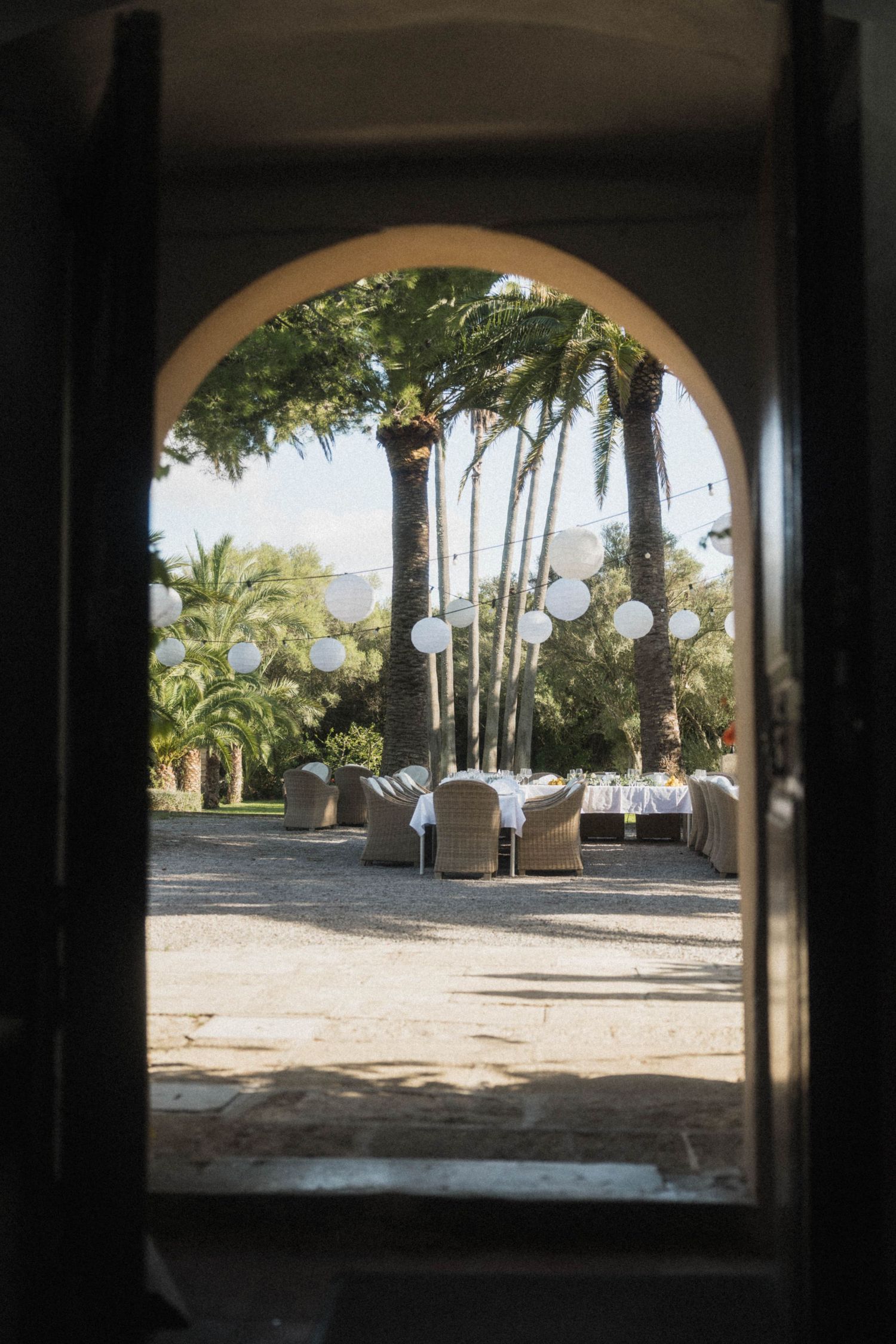 Arched doorway frames outdoor wedding reception with white lanterns and palm trees beyond.
