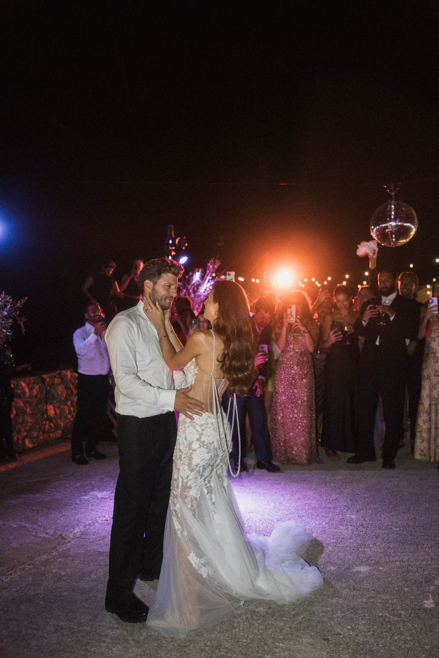 Bride in lace gown and groom share romantic first dance under disco ball at outdoor evening wedding.