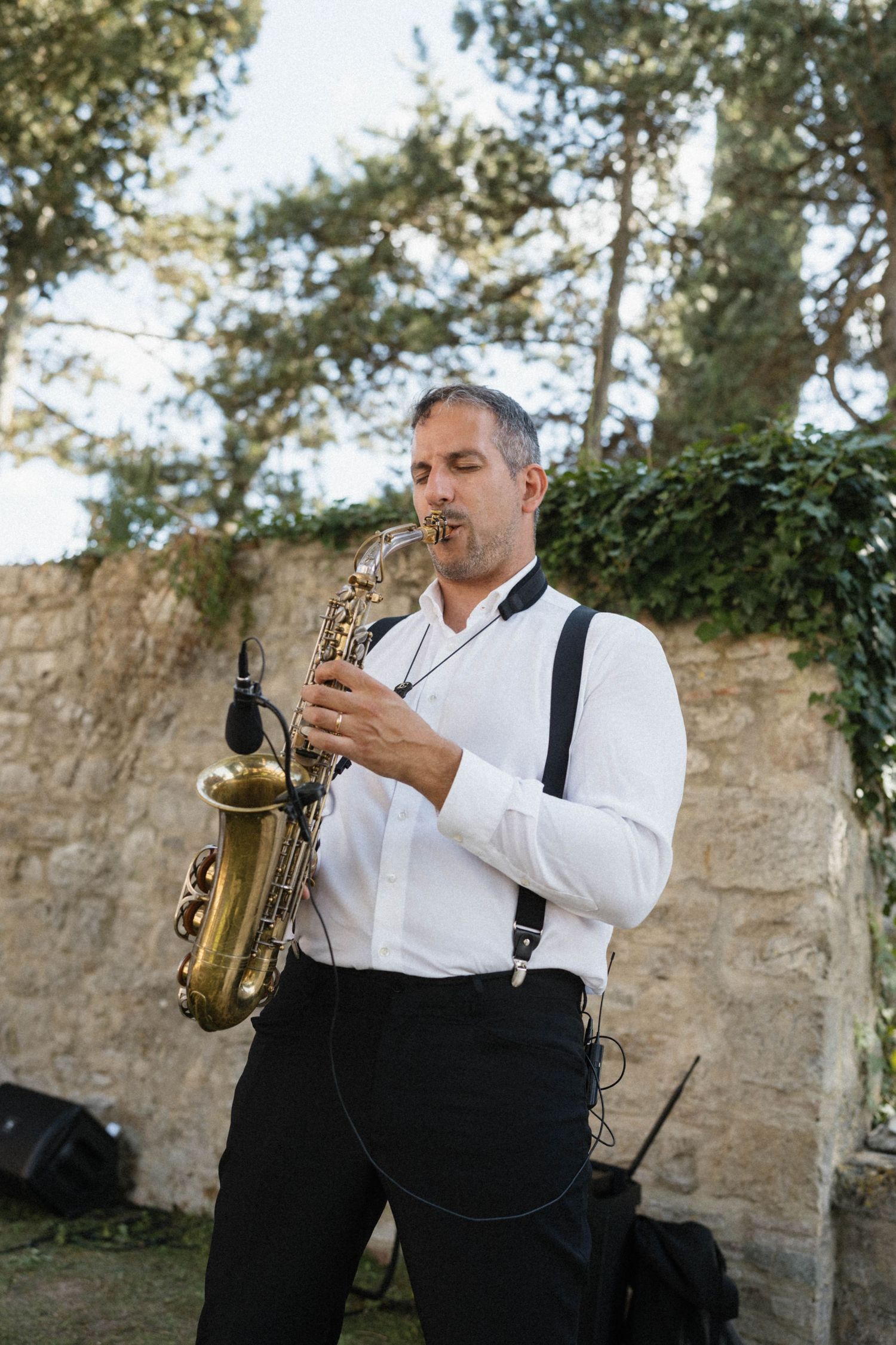 Saxophonist in white shirt and suspenders performing outdoors at an elegant wedding reception.