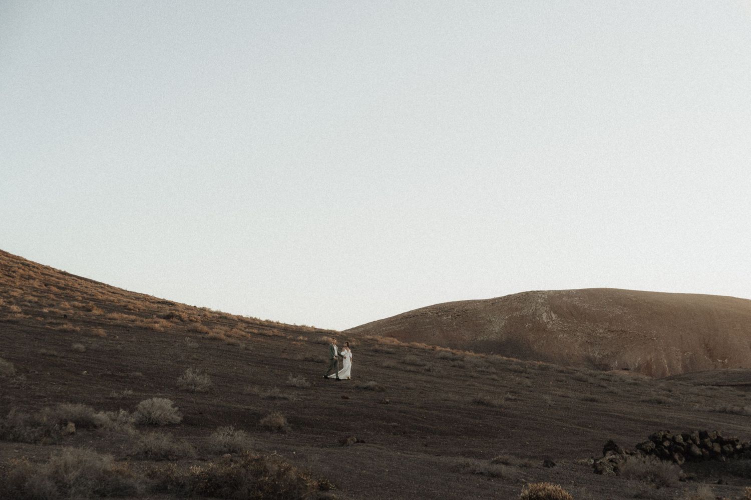 Couple in white wedding attire standing on a vast volcanic hillside landscape under a clear sky.
