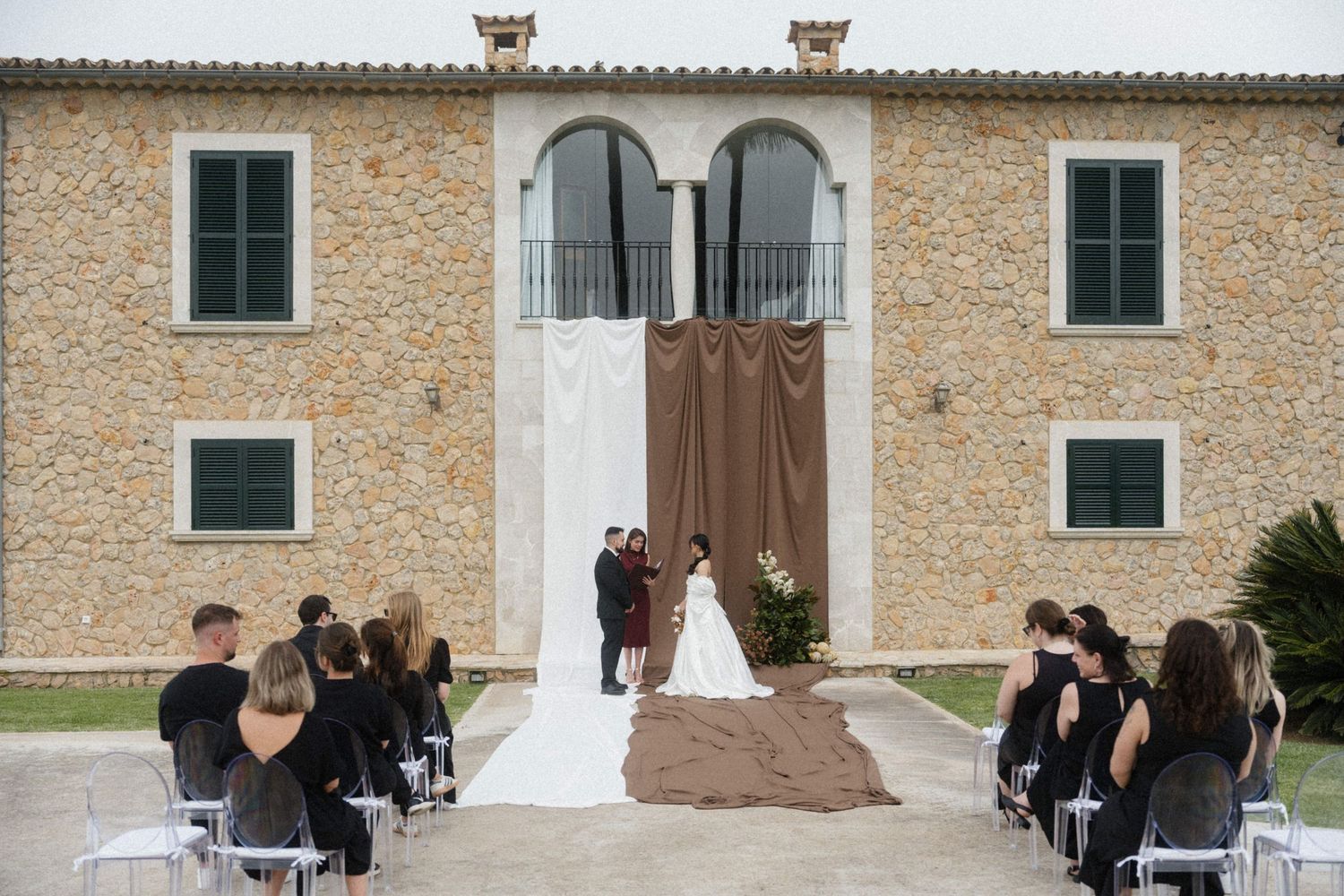 Outdoor wedding ceremony at a rustic stone villa with guests seated in white chairs.