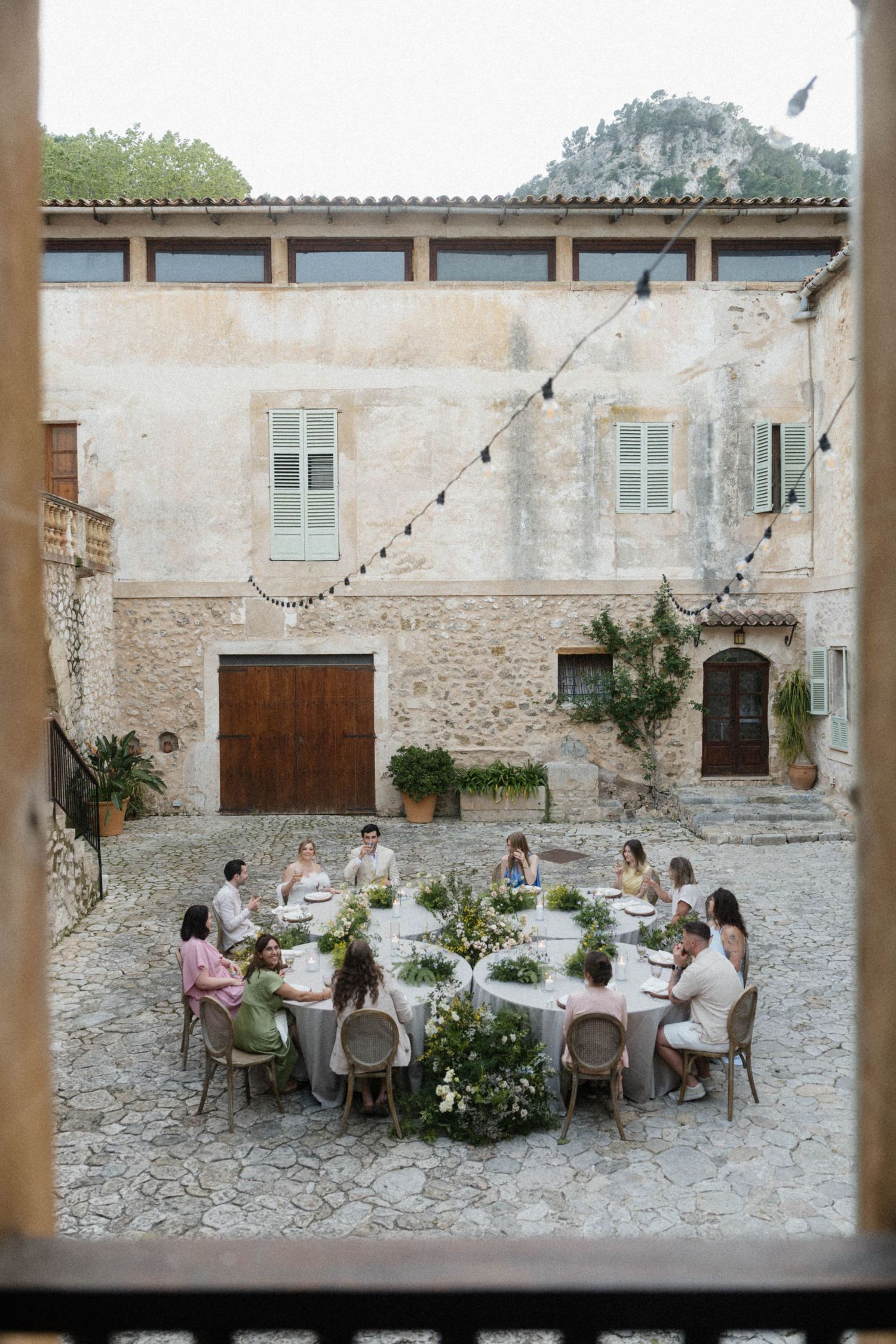 Wedding guests dining at round table in rustic stone courtyard with string lights overhead.