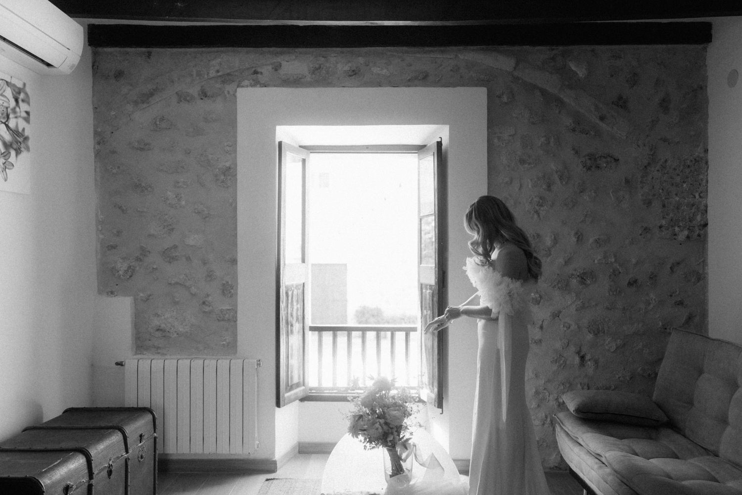 Black and white photo of bride in elegant wedding dress standing by open door in rustic stone room.