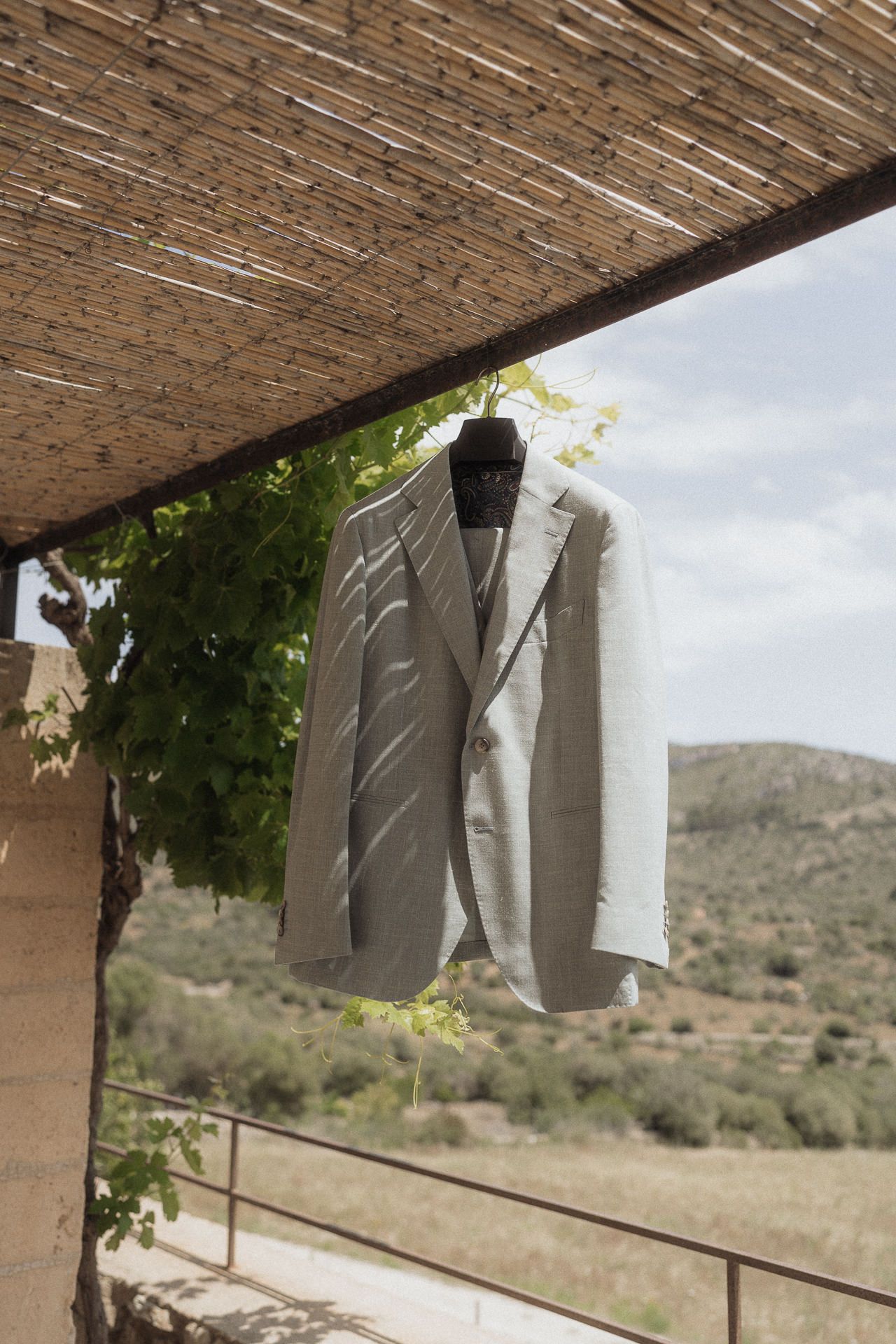 Light gray suit jacket hanging on a hanger under a bamboo pergola with scenic hills in the background.