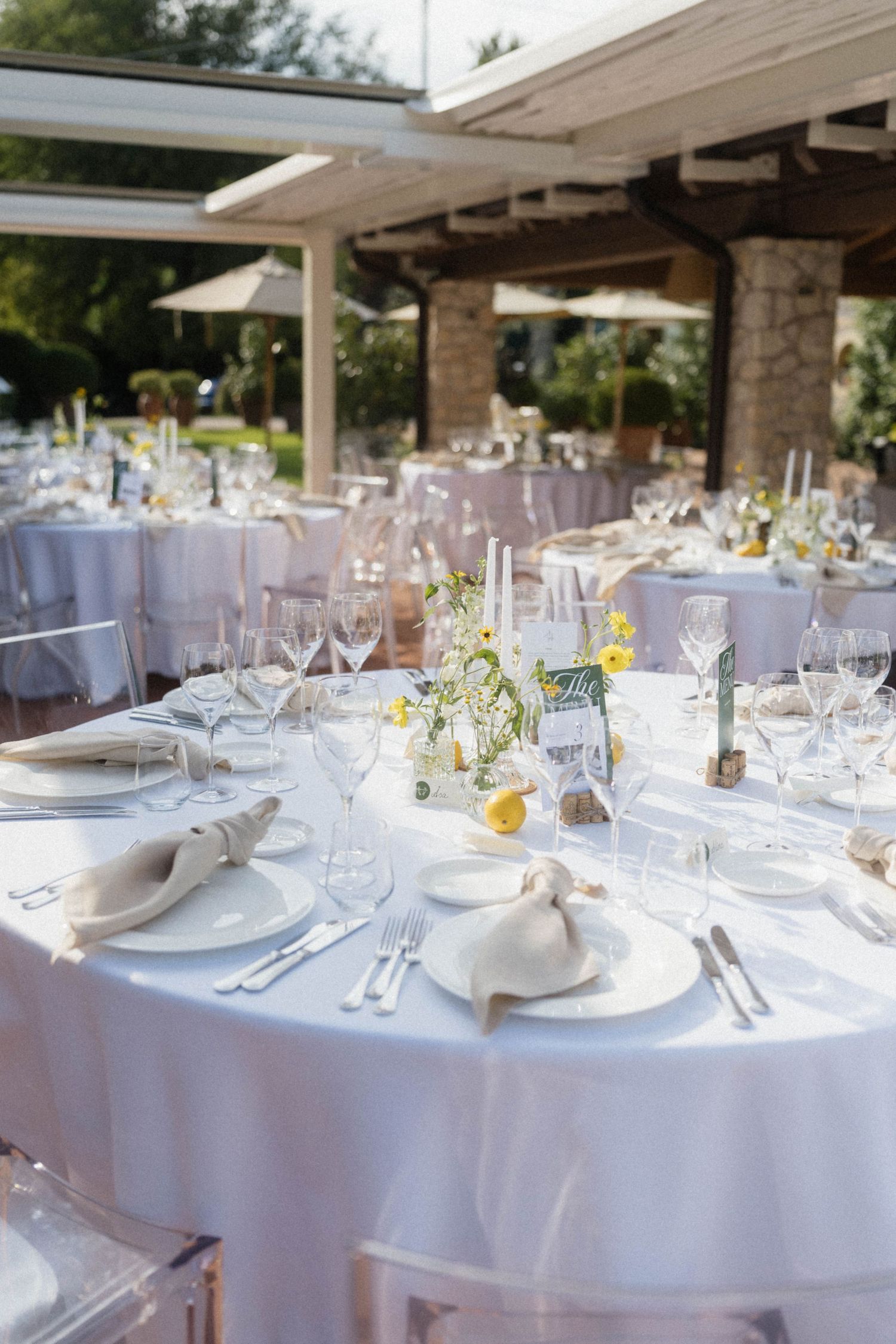 Elegant outdoor wedding reception with white round tables, crystal glasses, floral centerpieces, and ghost chairs under a pergola.