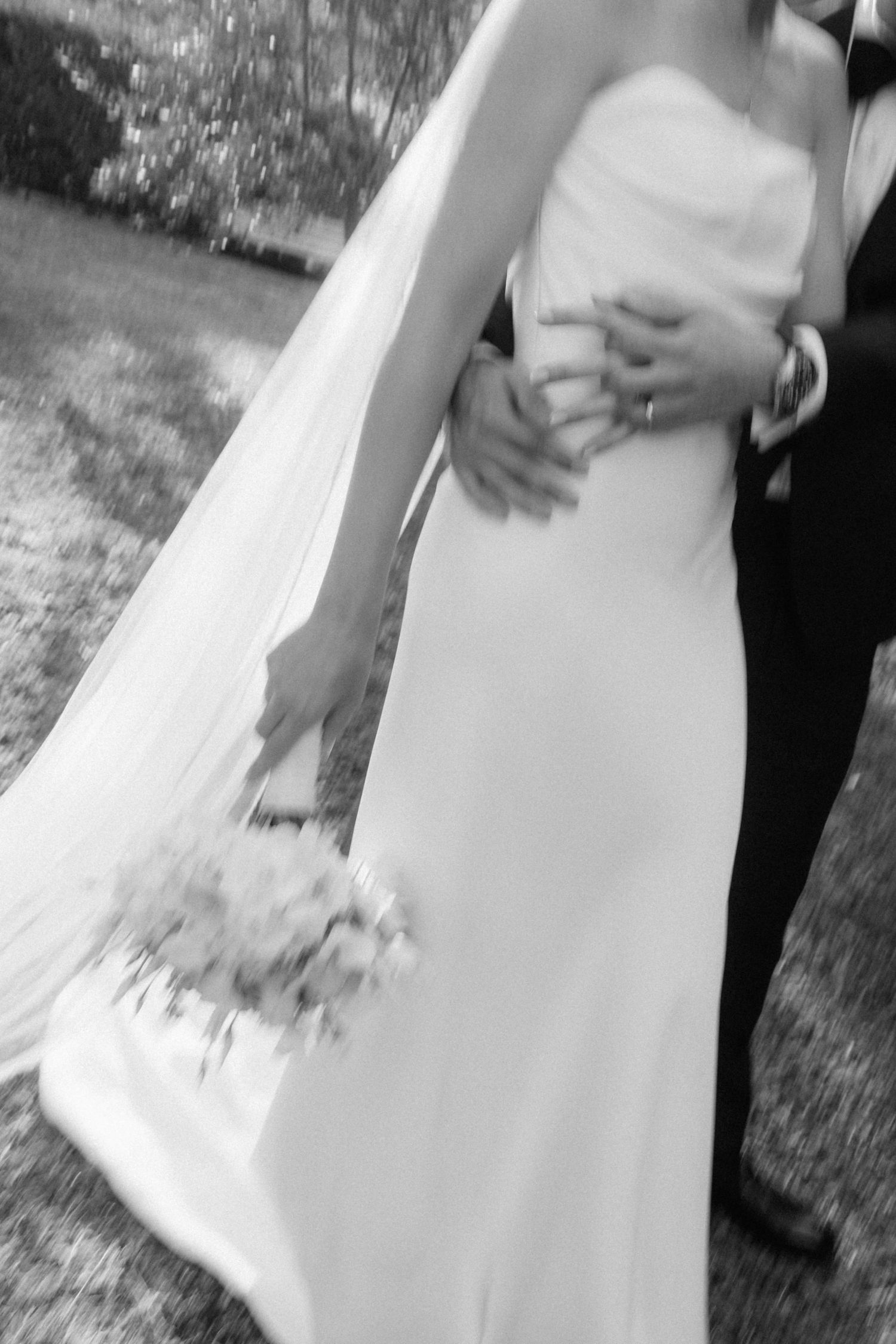 Black and white artistic motion blur of bride holding bouquet with groom embracing her waist outdoors.