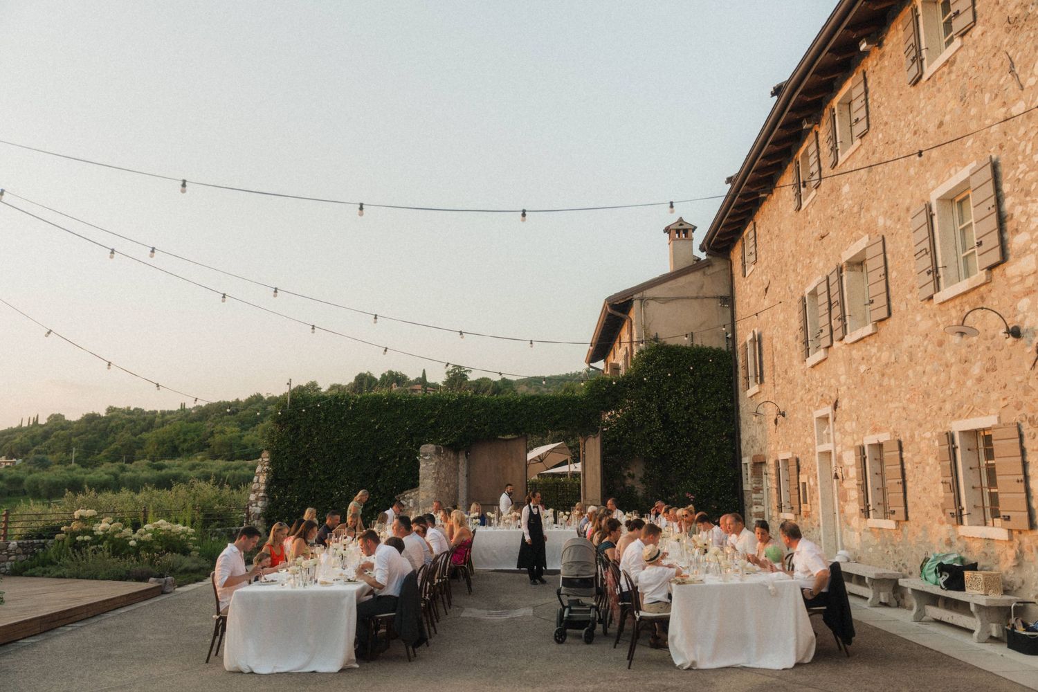 Guests dine at long white tables during an outdoor wedding reception at a rustic stone villa at dusk.