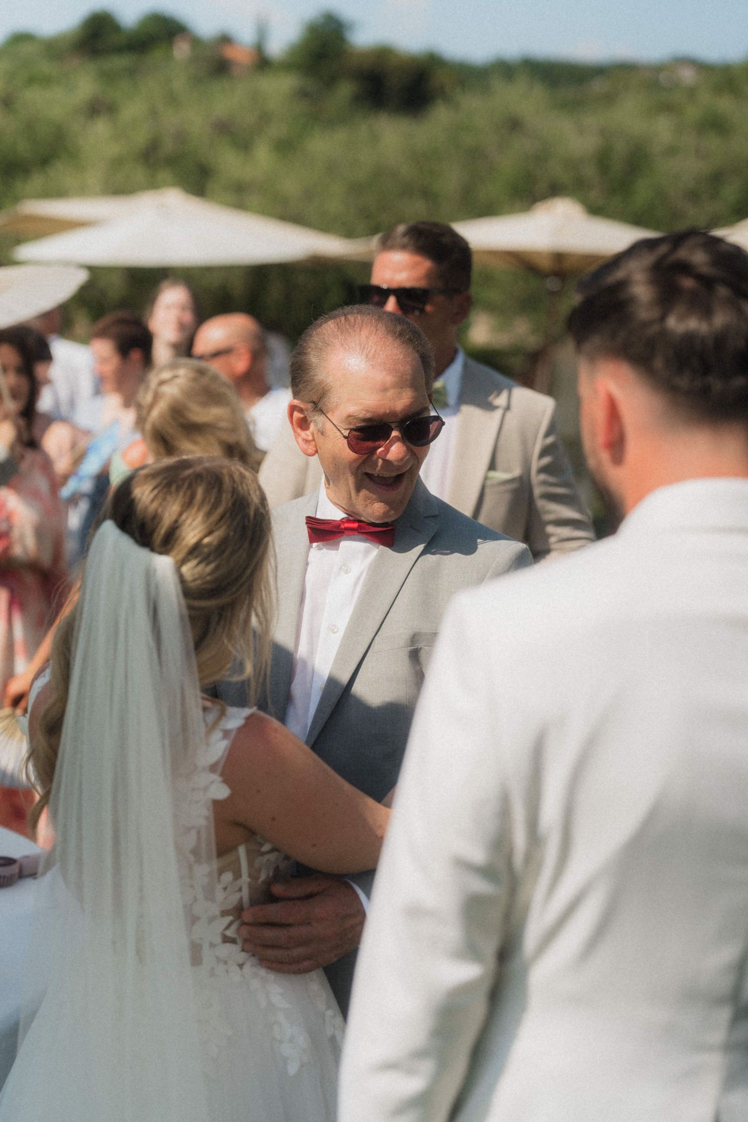 Father of the bride smiling warmly in red bow tie during outdoor wedding ceremony.