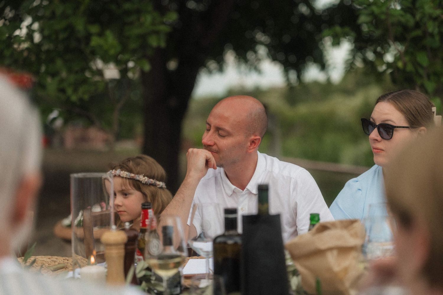 Wedding guests including a child with floral crown seated at outdoor reception dinner table.