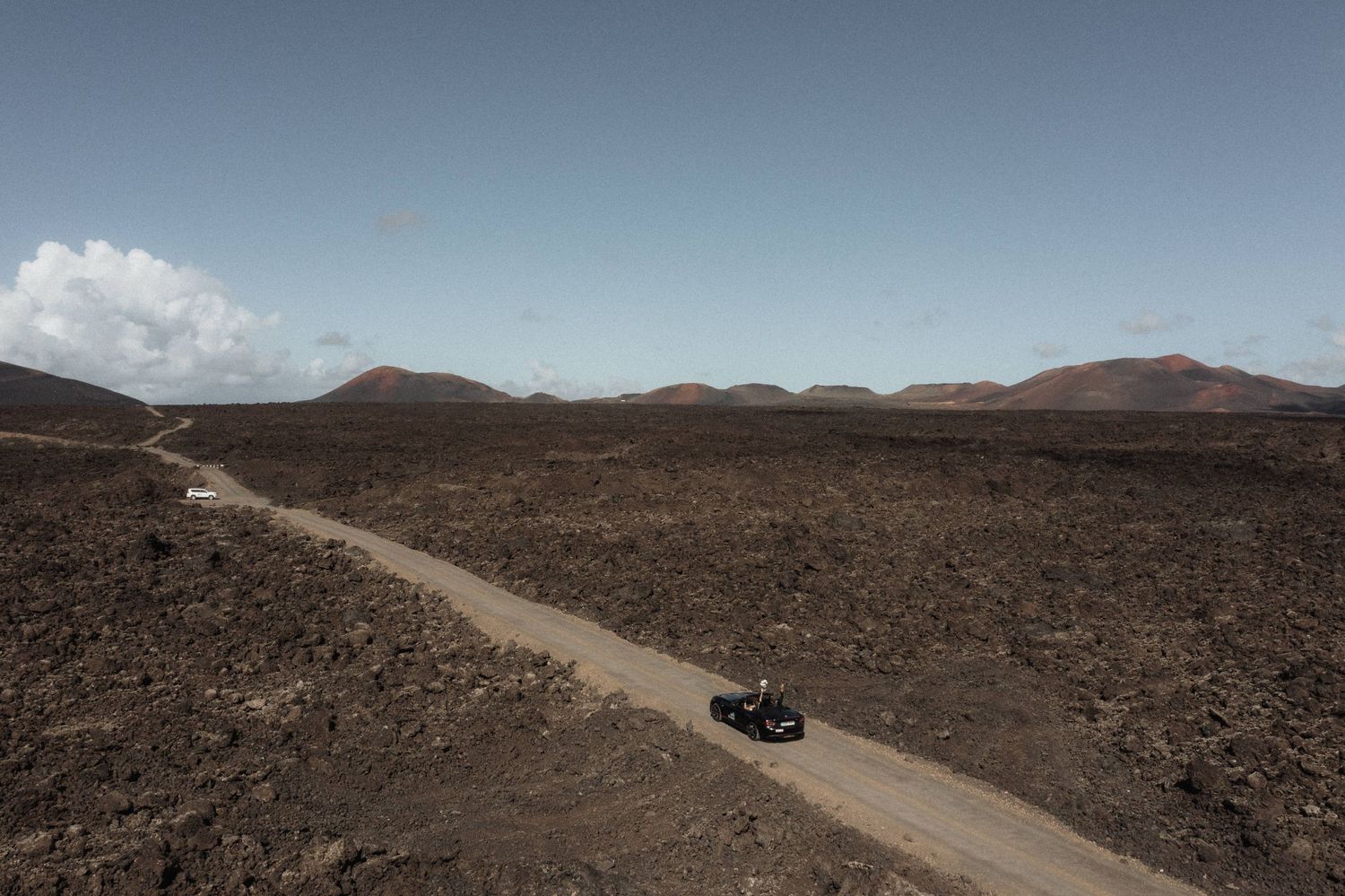Aerial view of a vehicle driving on a remote dirt road through dramatic volcanic lava landscape.