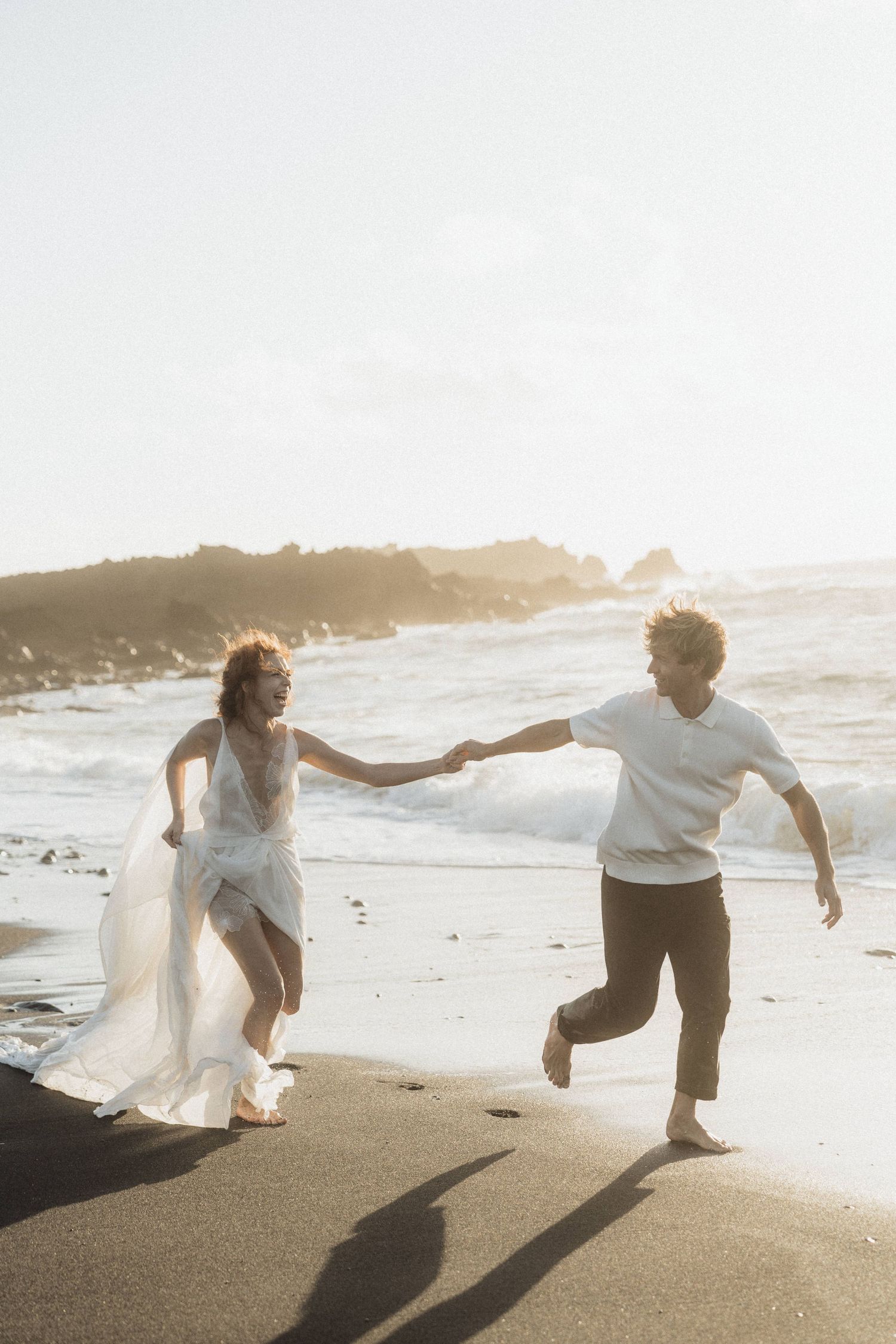Couple runs joyfully hand in hand along black sand beach in golden sunlight.