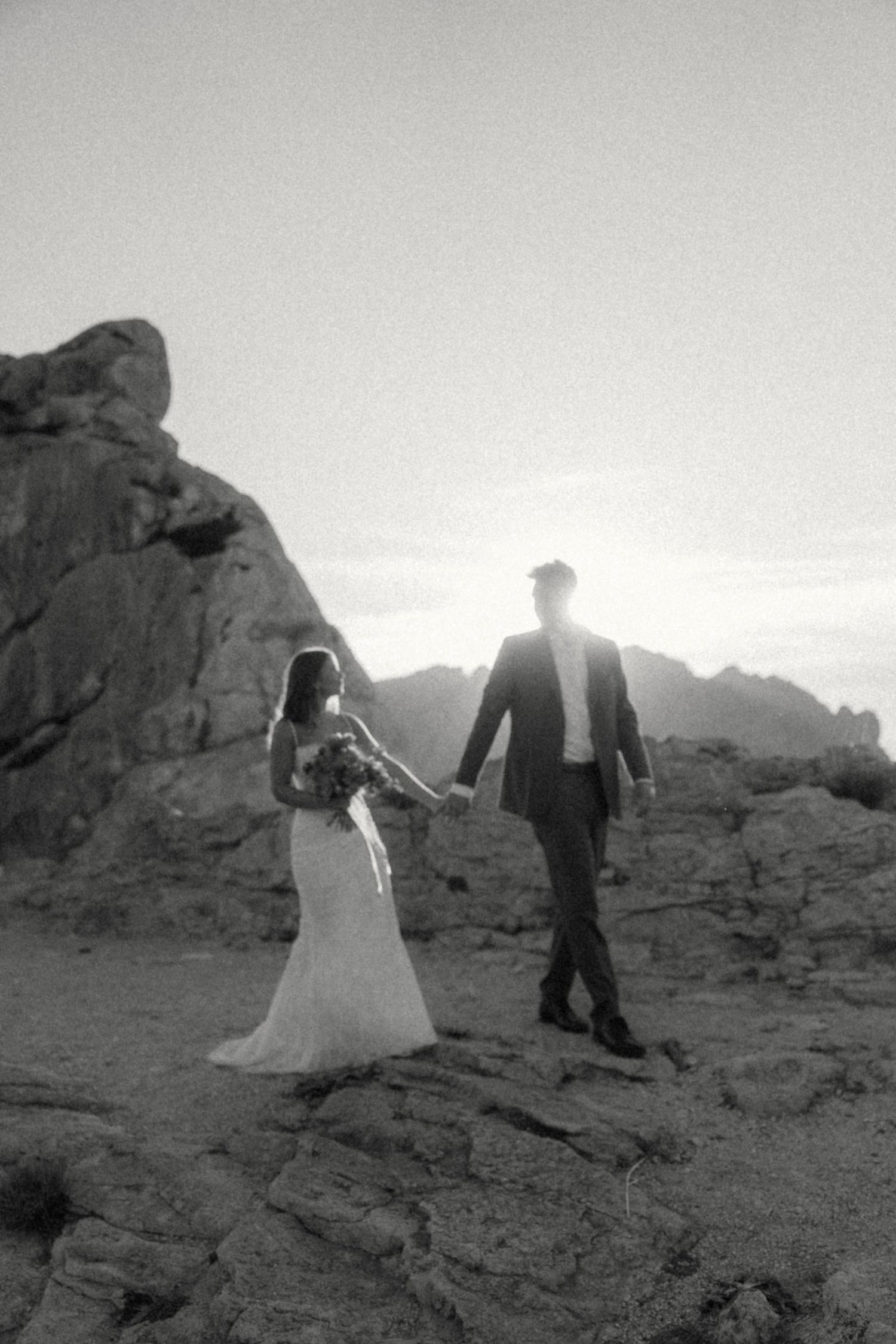 Black and white photo of bride and groom holding hands on rocky terrain with dramatic backlit sunset behind them.