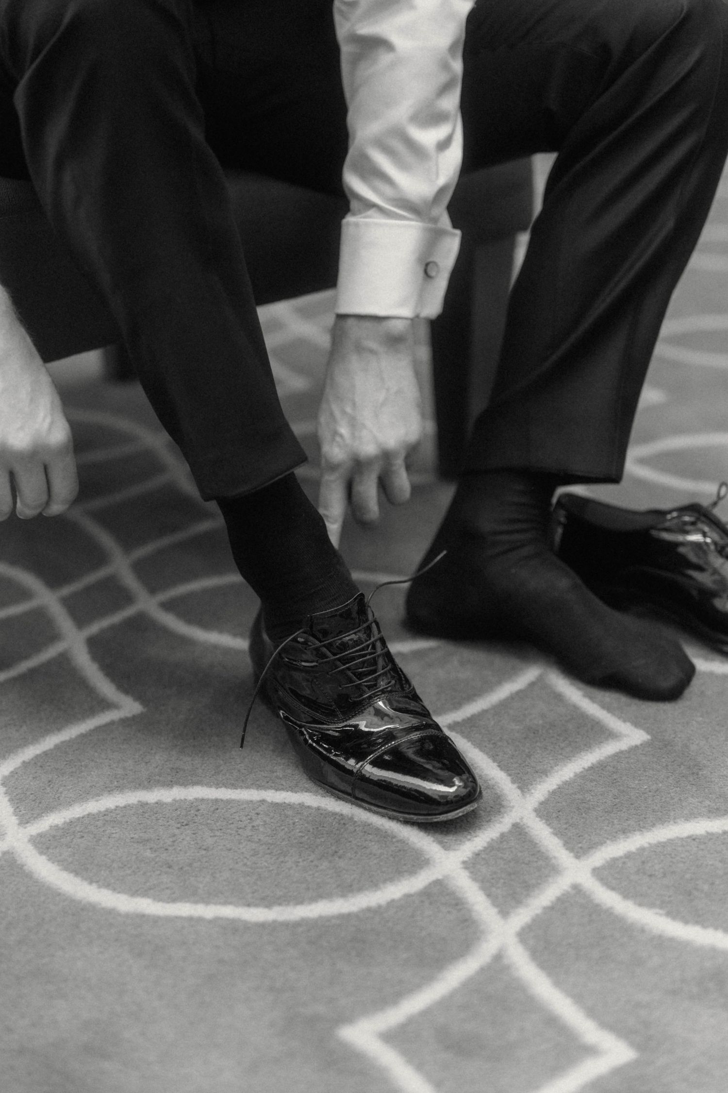 Groom tying his black patent leather dress shoes while seated, wearing dark suit trousers and black socks.