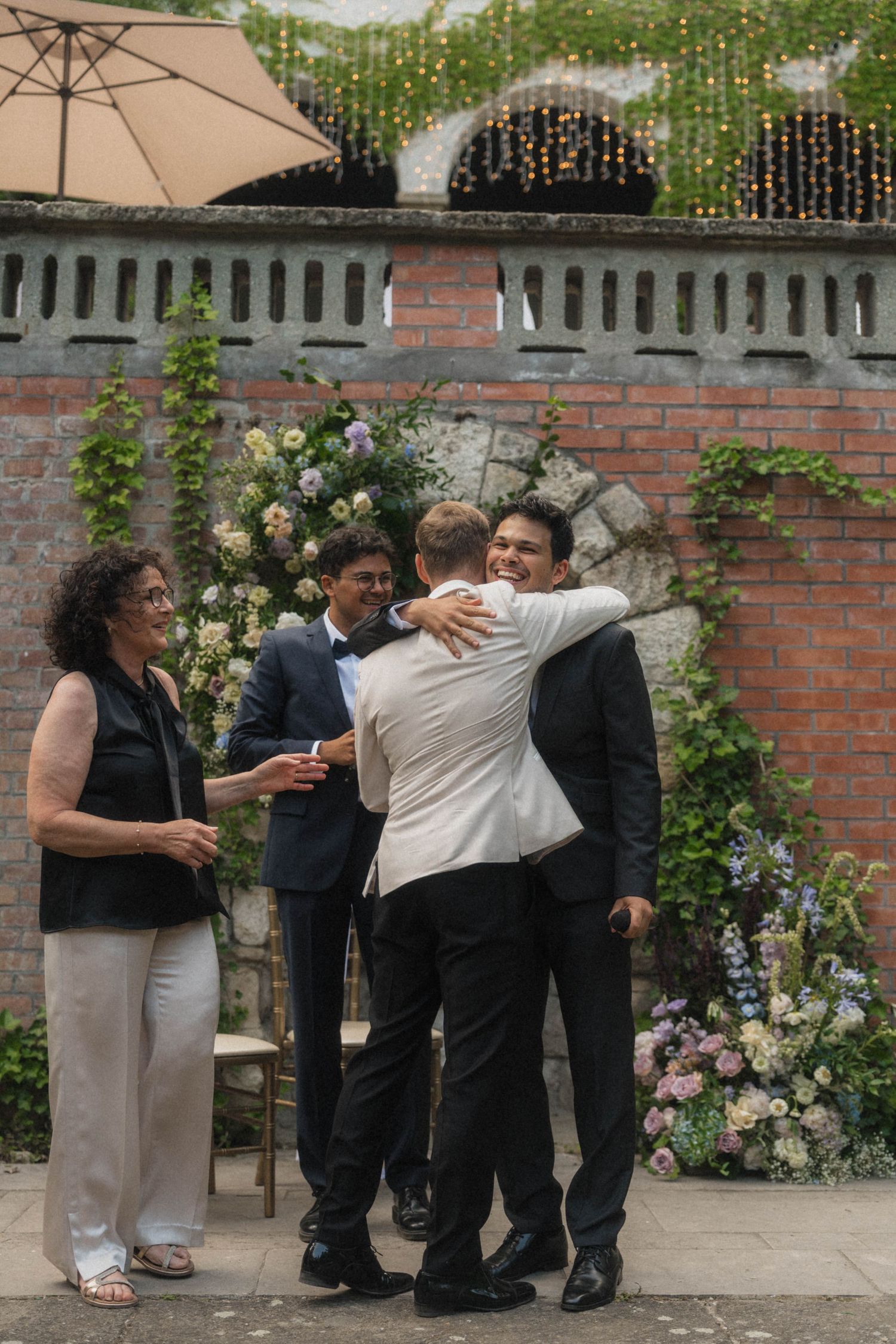 Groom embracing a guest at an outdoor wedding ceremony with floral arch and brick wall backdrop.