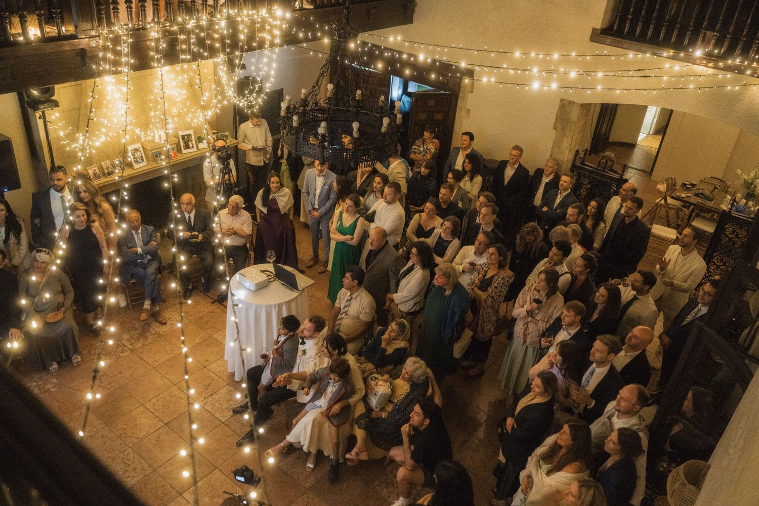 Aerial view of wedding reception guests gathered under warm string lights inside a rustic venue.