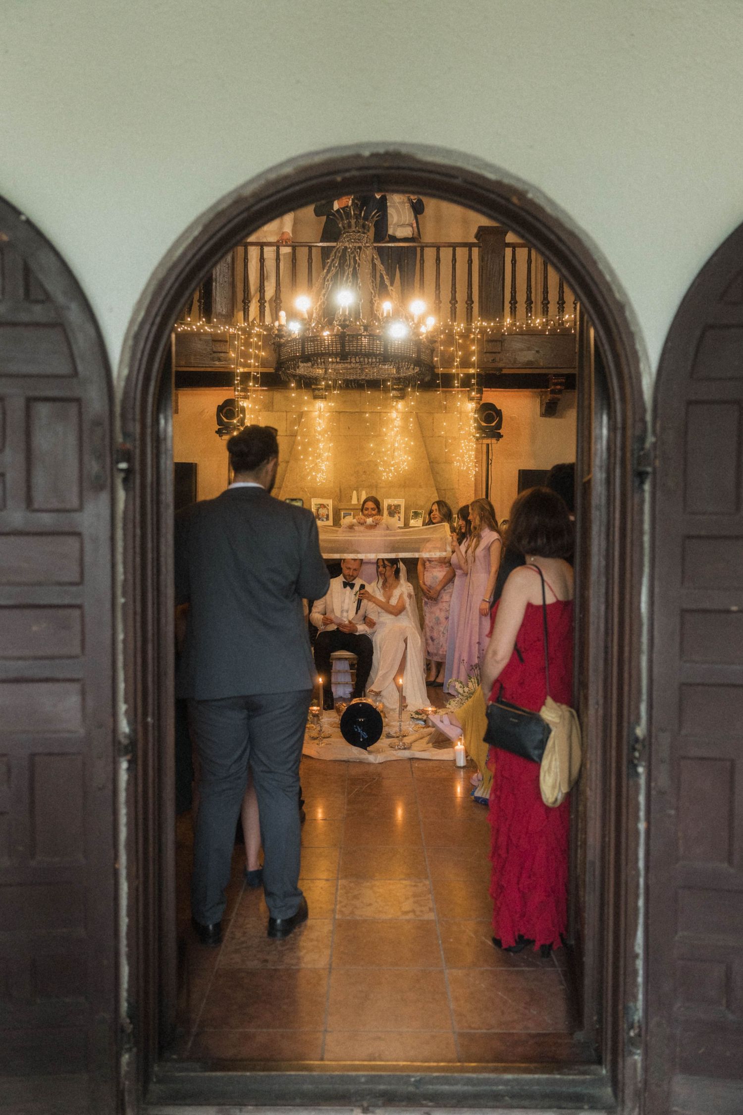 Wedding guests gathered in warmly lit historic venue viewed through ornate arched wooden doorway with fairy lights.