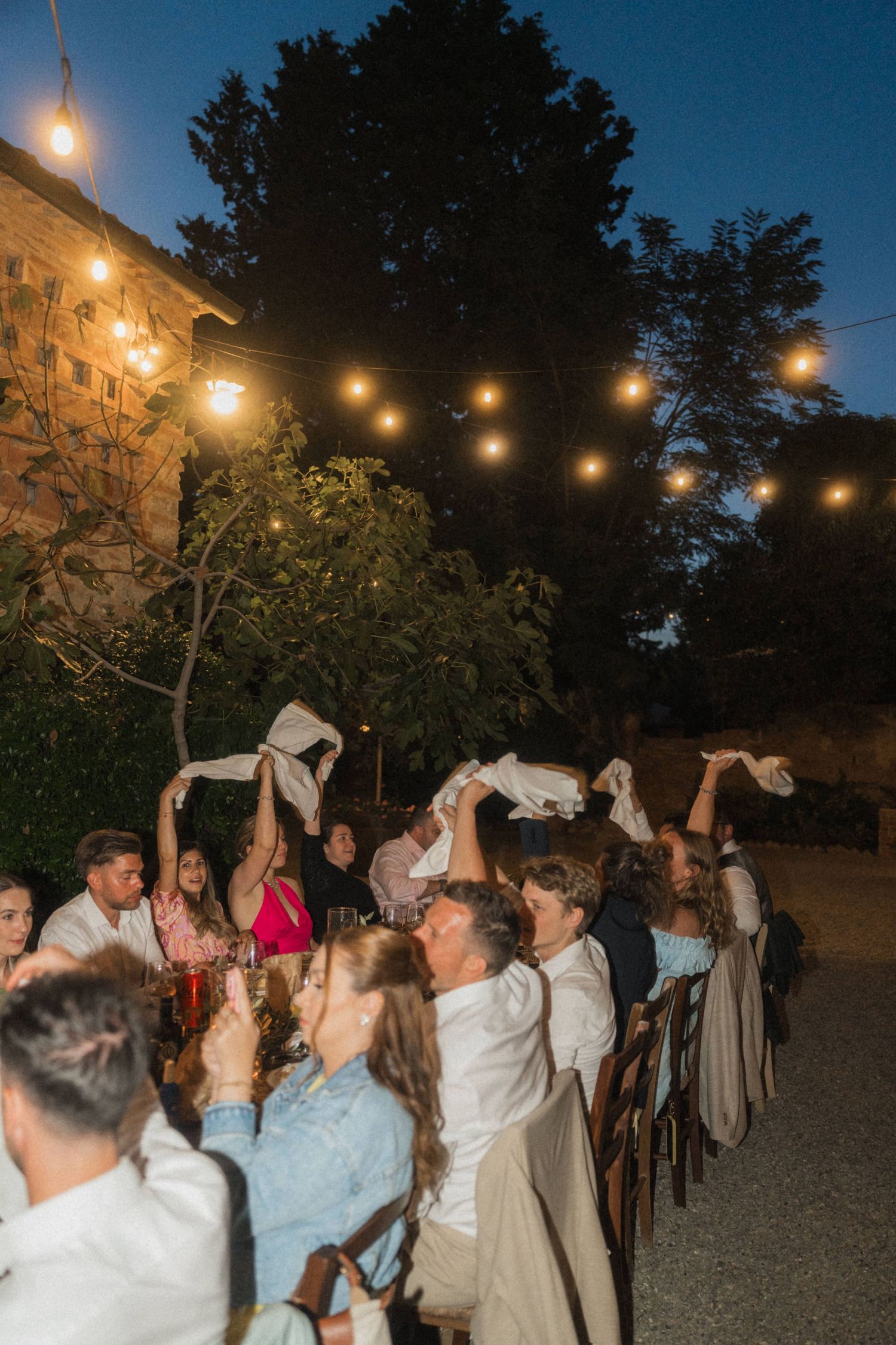 Wedding guests waving white napkins at outdoor evening reception under warm string lights.