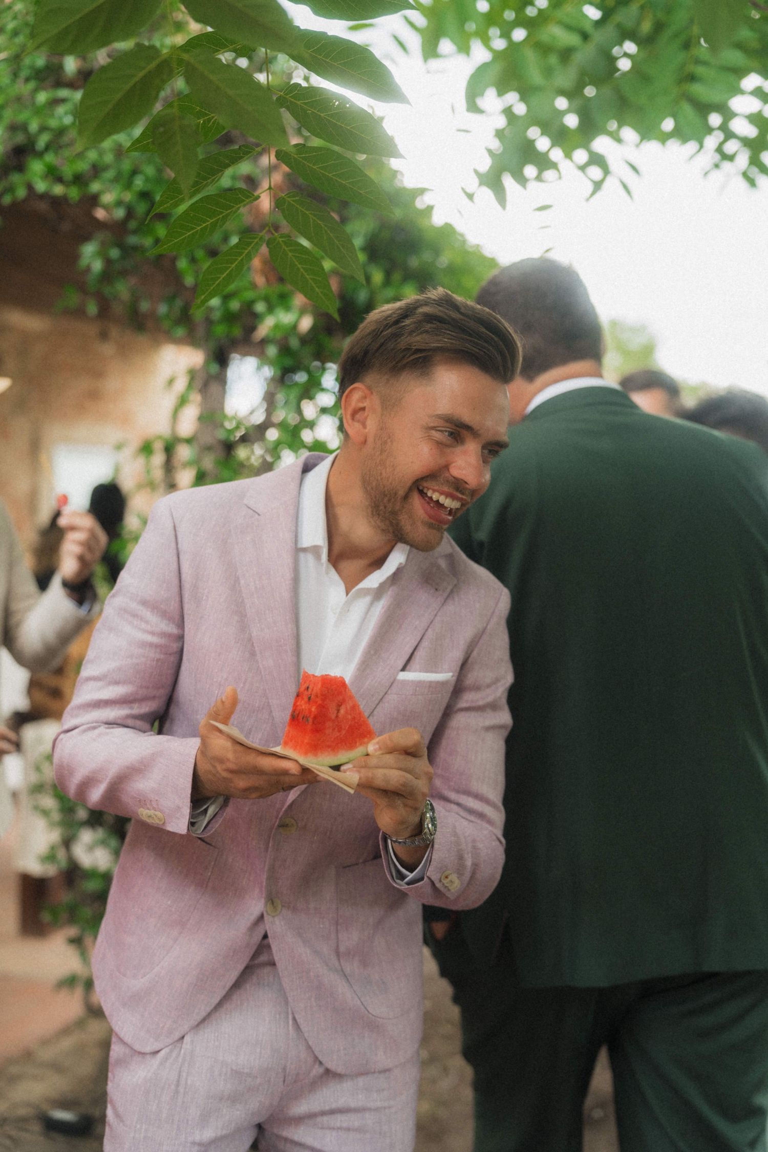Smiling man in pink suit holding watermelon slice at outdoor wedding reception.