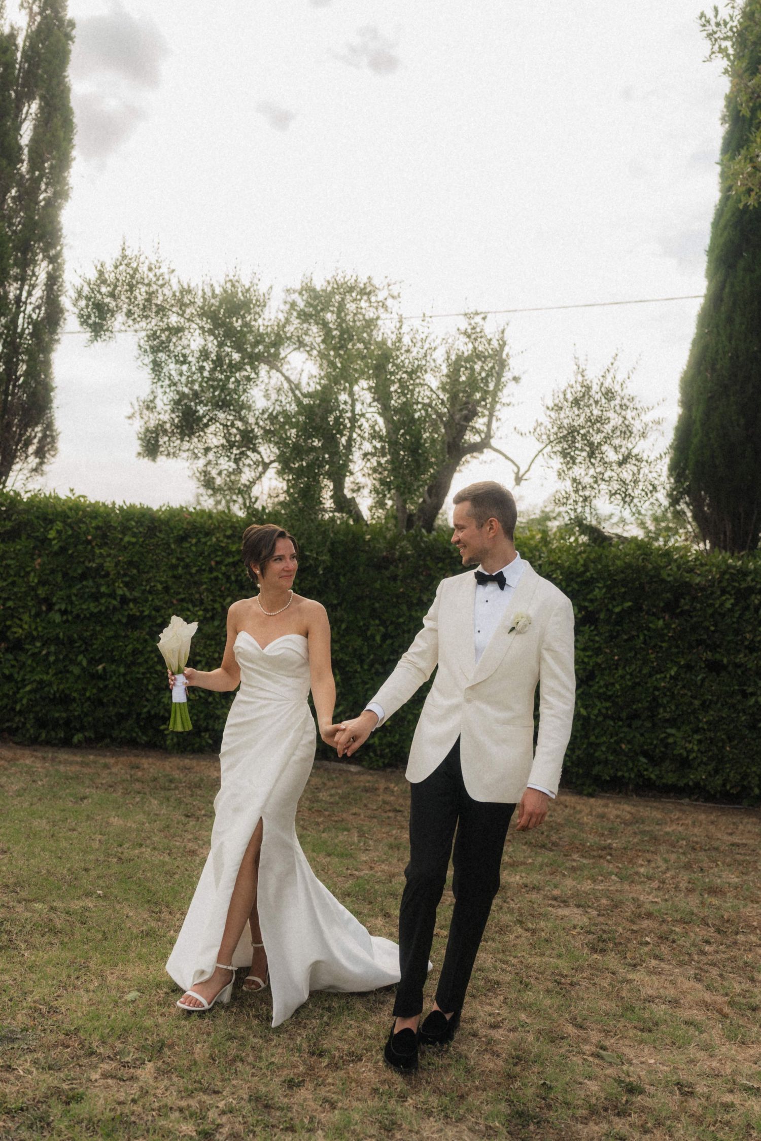 Bride and groom holding hands walking in garden with cypress trees on wedding day.