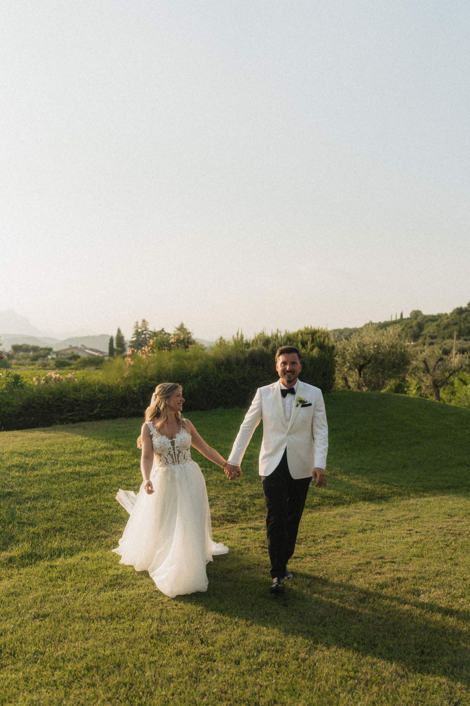 Bride and groom holding hands walking on green lawn at sunset.