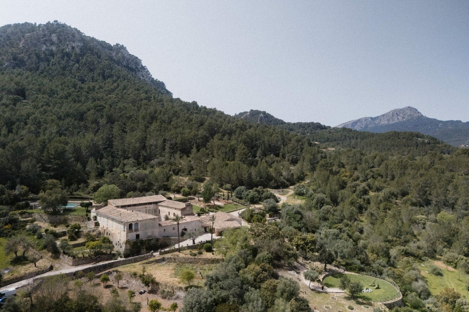 Aerial view of a historic stone estate nestled among lush green trees with dramatic rocky mountains in the background.