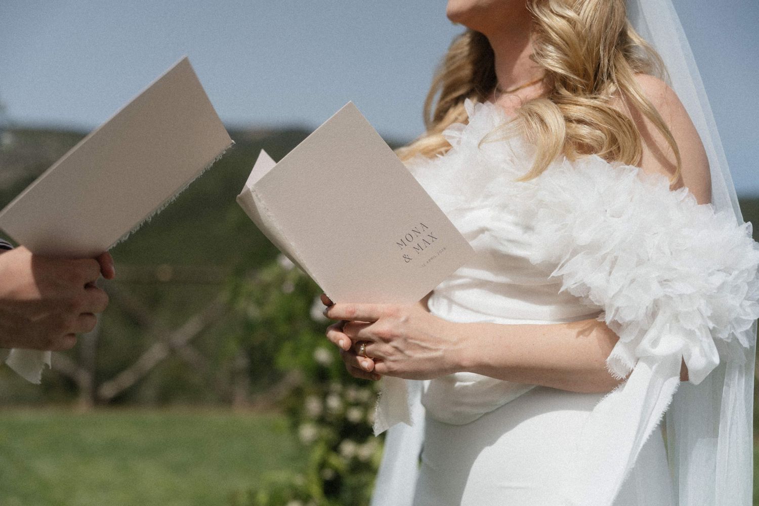 Bride in a white feathered wedding dress holding an open ceremony booklet during an outdoor wedding ceremony.
