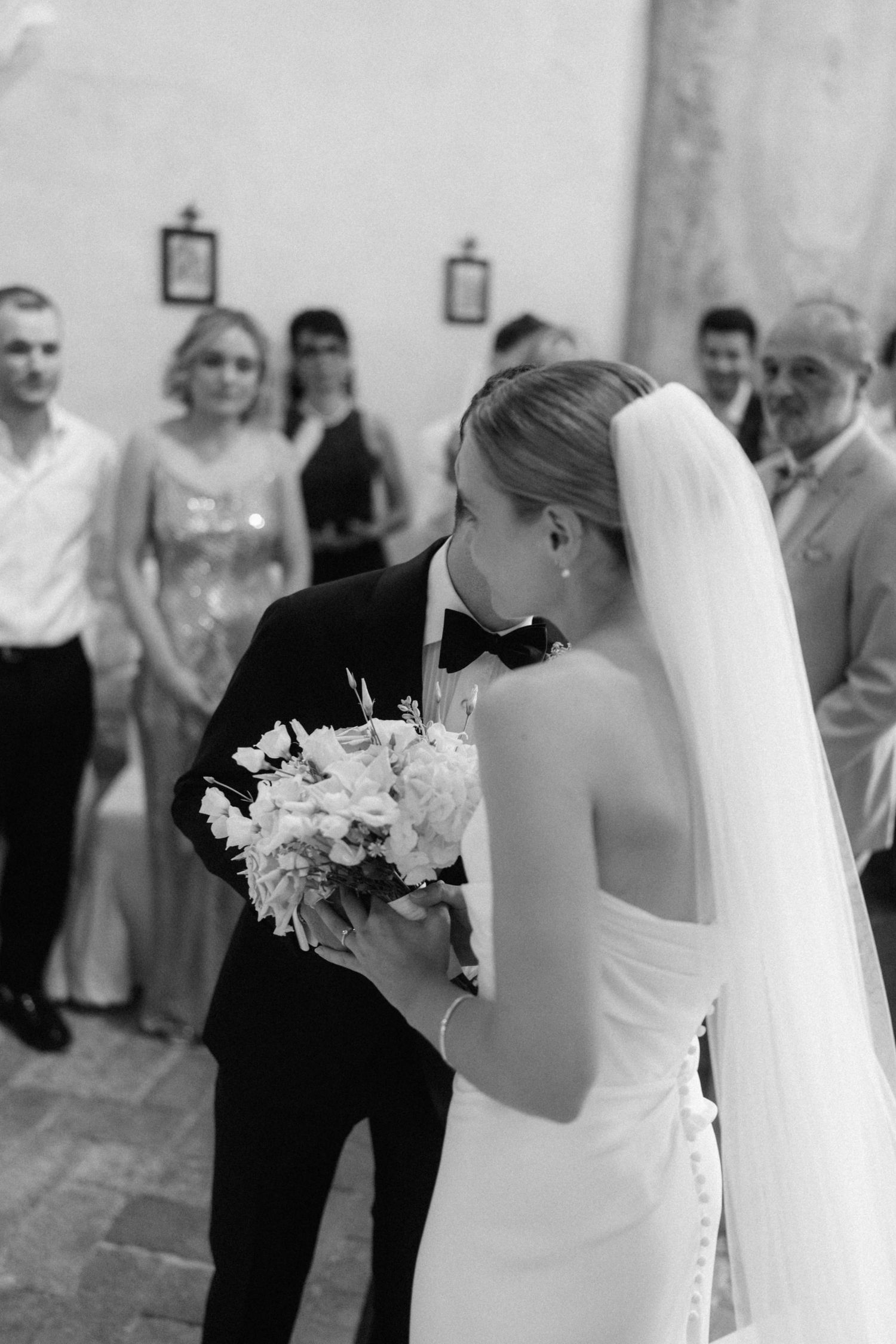 Black and white wedding photo of bride and groom kissing at altar with bouquet, guests watching in background.