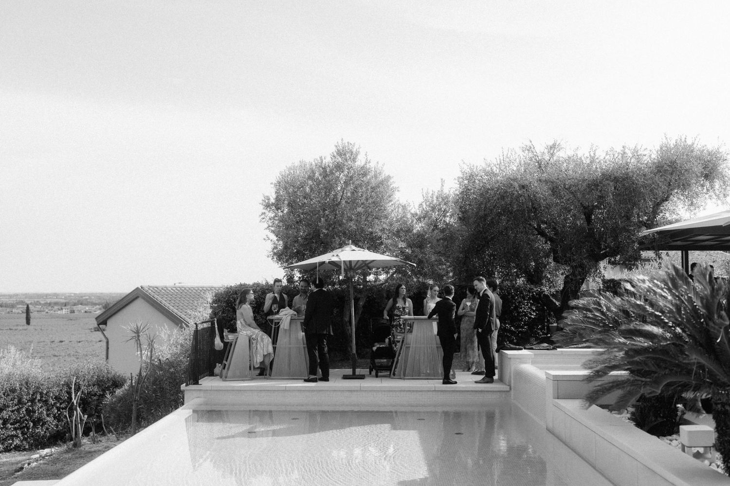 Black and white outdoor wedding ceremony with guests gathered near a pool, olive trees, and umbrella at a elegant venue.